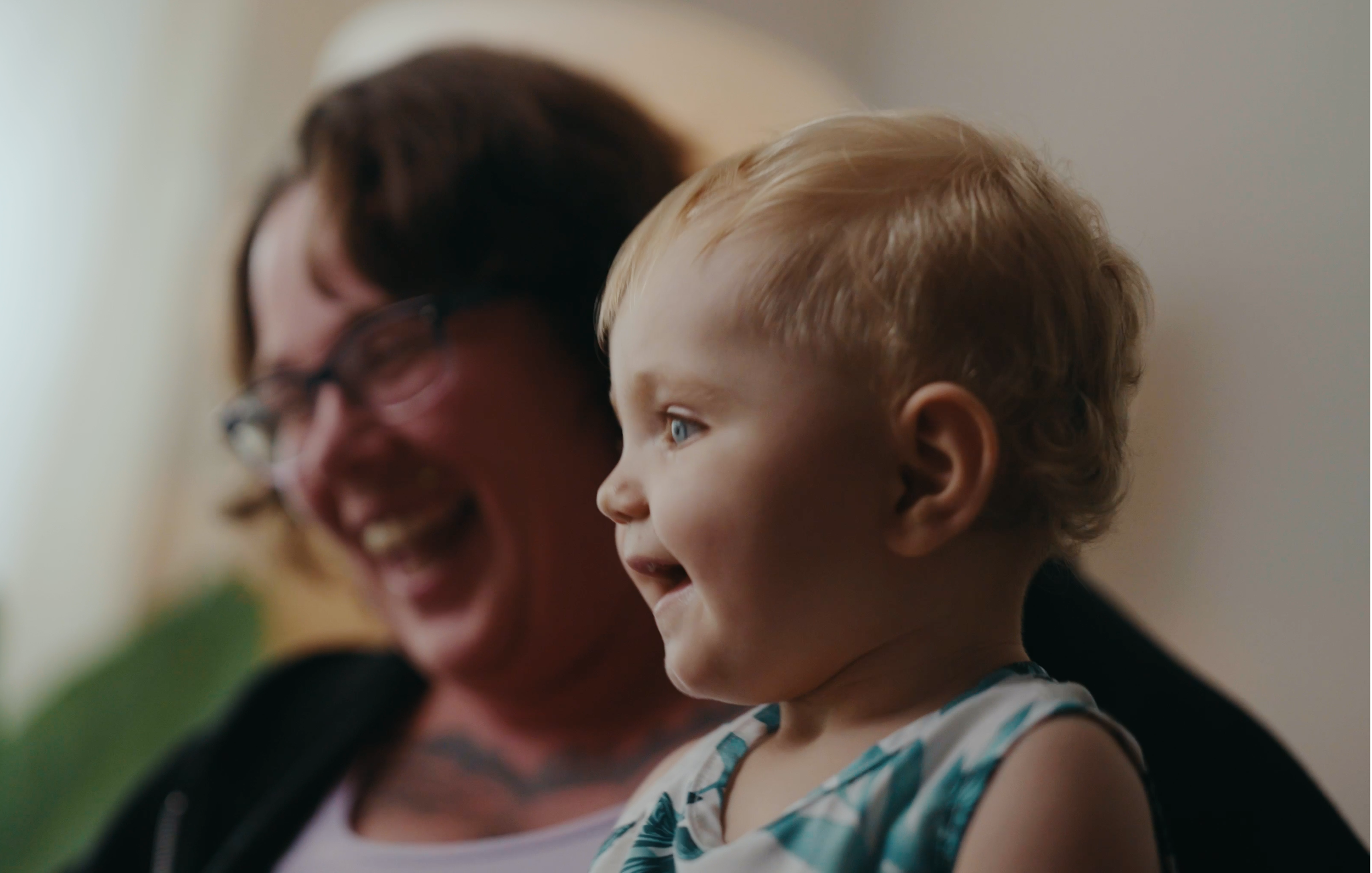 A young child with blond hair and blue eyes smiling while sitting on an adult woman's lap. The woman, wearing glasses and a white cap, is smiling and appears to be laughing.