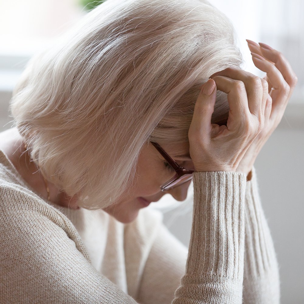 An elderly woman with gray hair, wearing glasses and a beige sweater, sitting with her hand on her forehead.