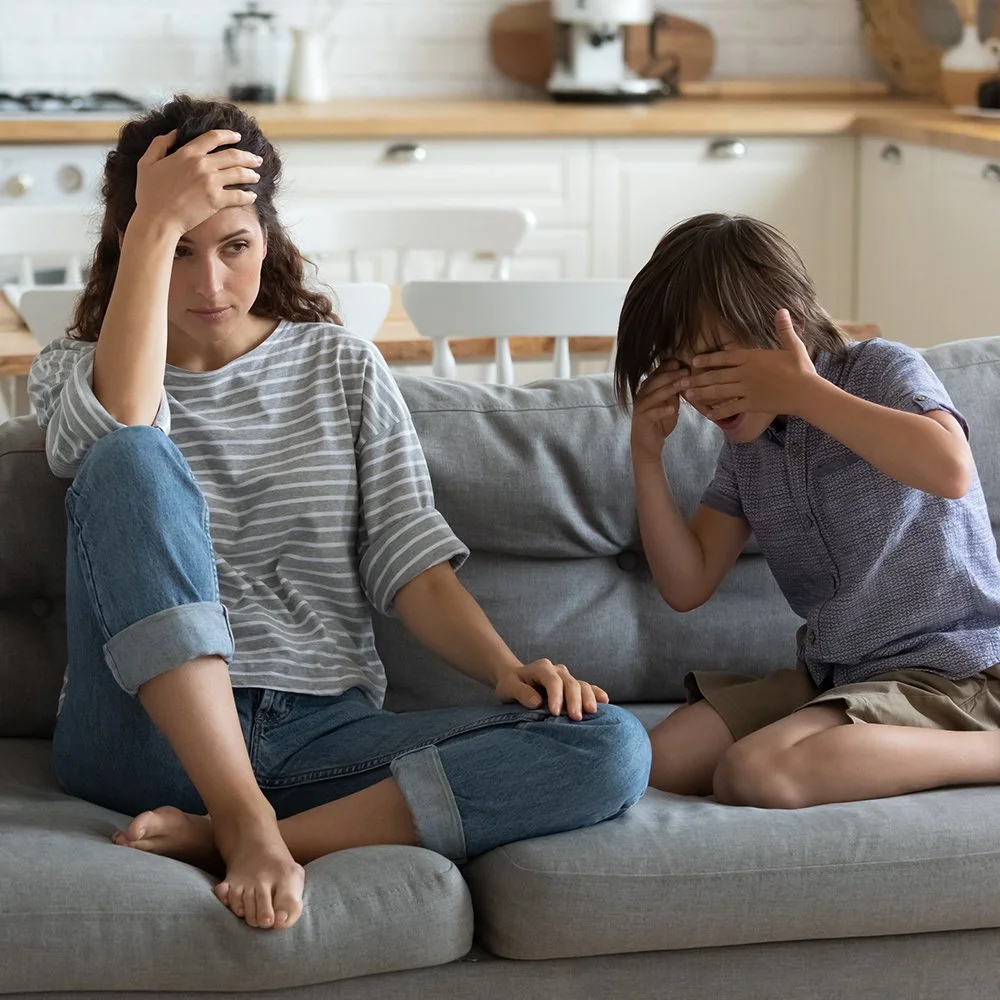 A woman and a young boy sitting on a gray couch in a kitchen, both touching their foreheads and faces with their hands, appearing distressed or upset.