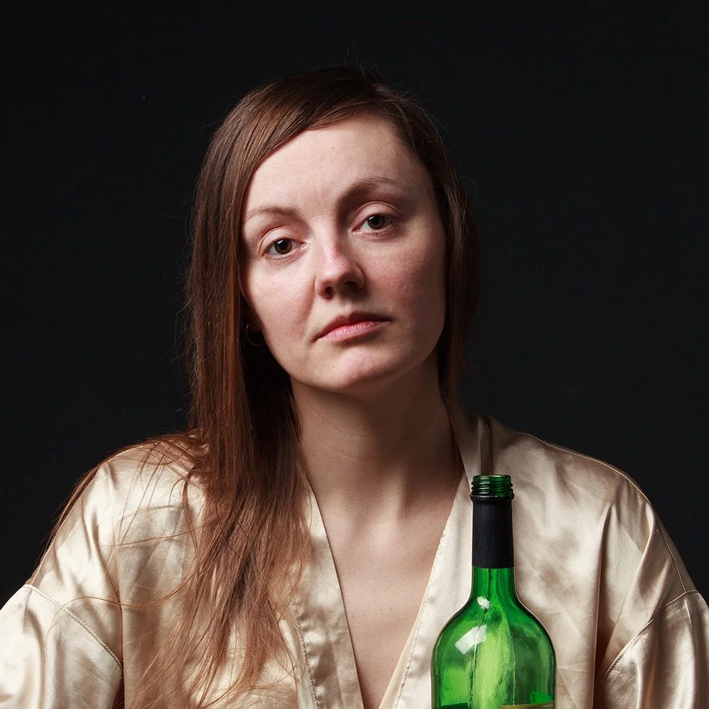 A woman with long brown hair and a neutral expression is holding a green bottle against a dark background.