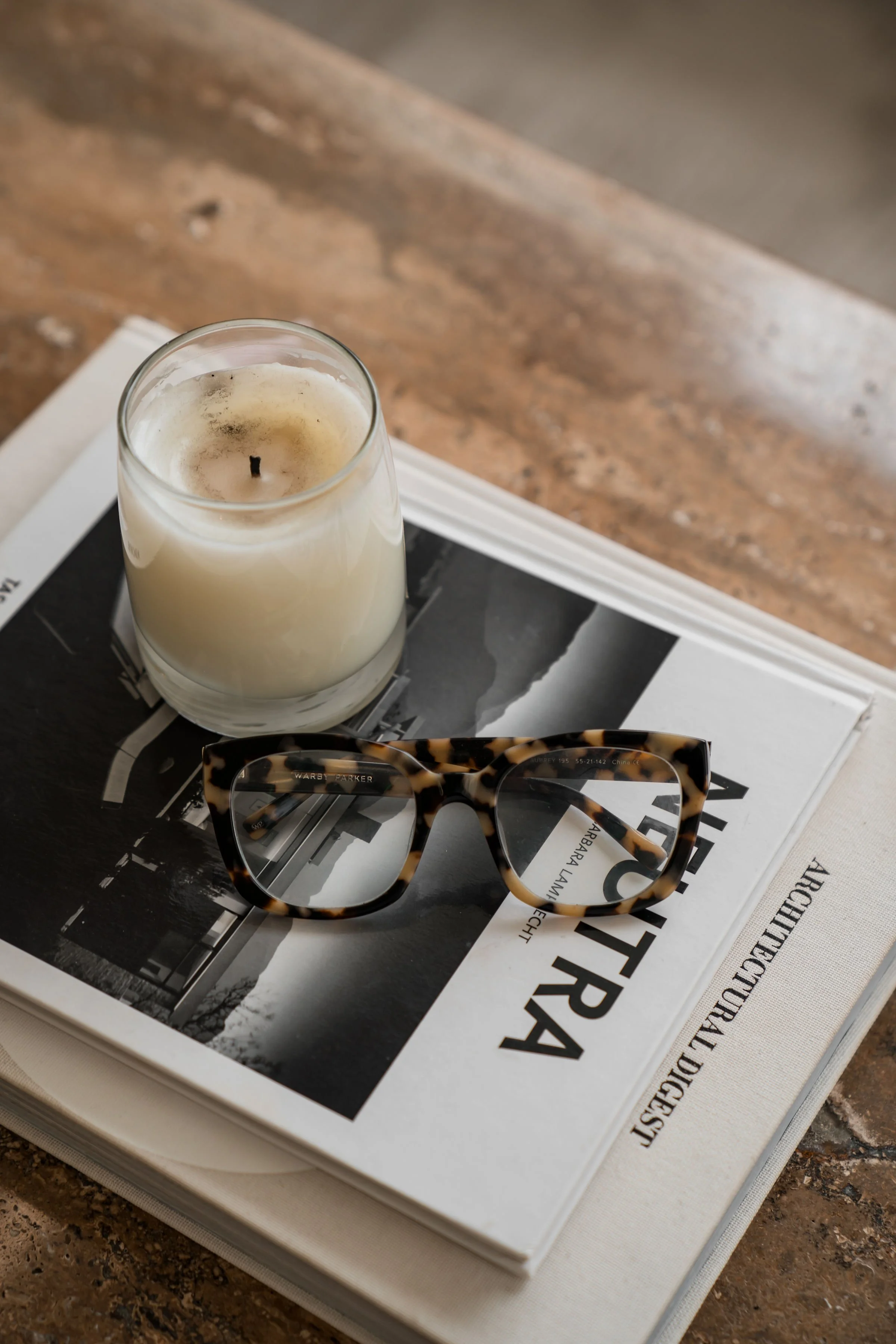 A pair of tortoiseshell eyeglasses resting on top of a magazine with a black and white photograph, placed on a white book or magazine on a granite countertop. A glass candle with a burning wick and melted wax is positioned behind the glasses.