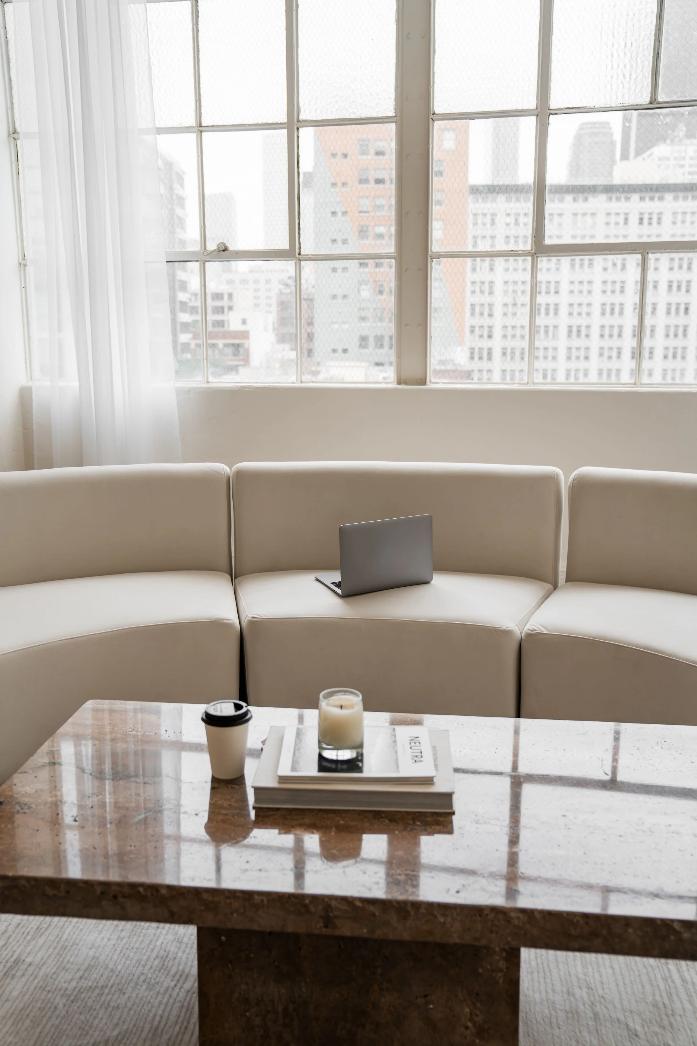Interior of a modern urban café or lounge with a white curved sofa facing large window panels, a marble coffee table with a coffee cup, a lit candle, and a magazine on it, city buildings outside the window.