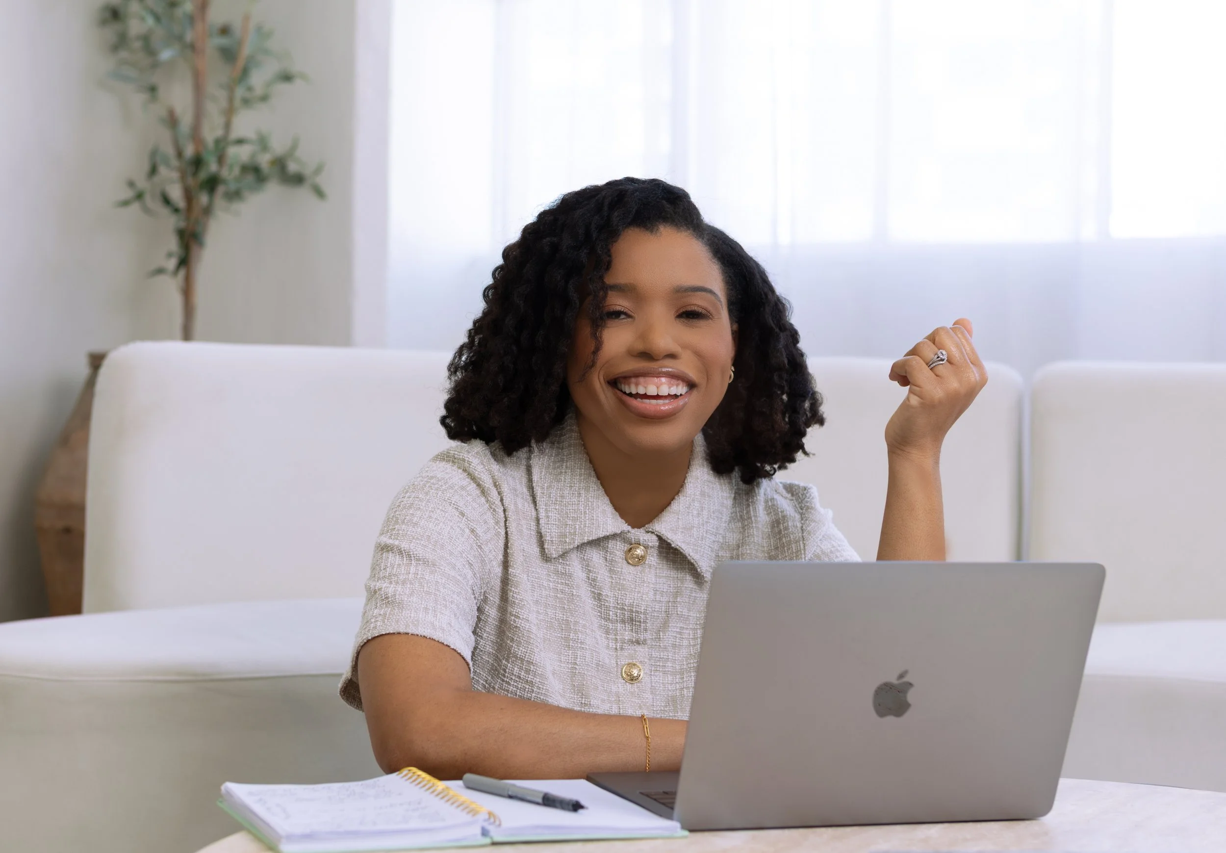 A woman smiling and raising her fist in front of a laptop in a bright room, with a notebook and pen on the table.