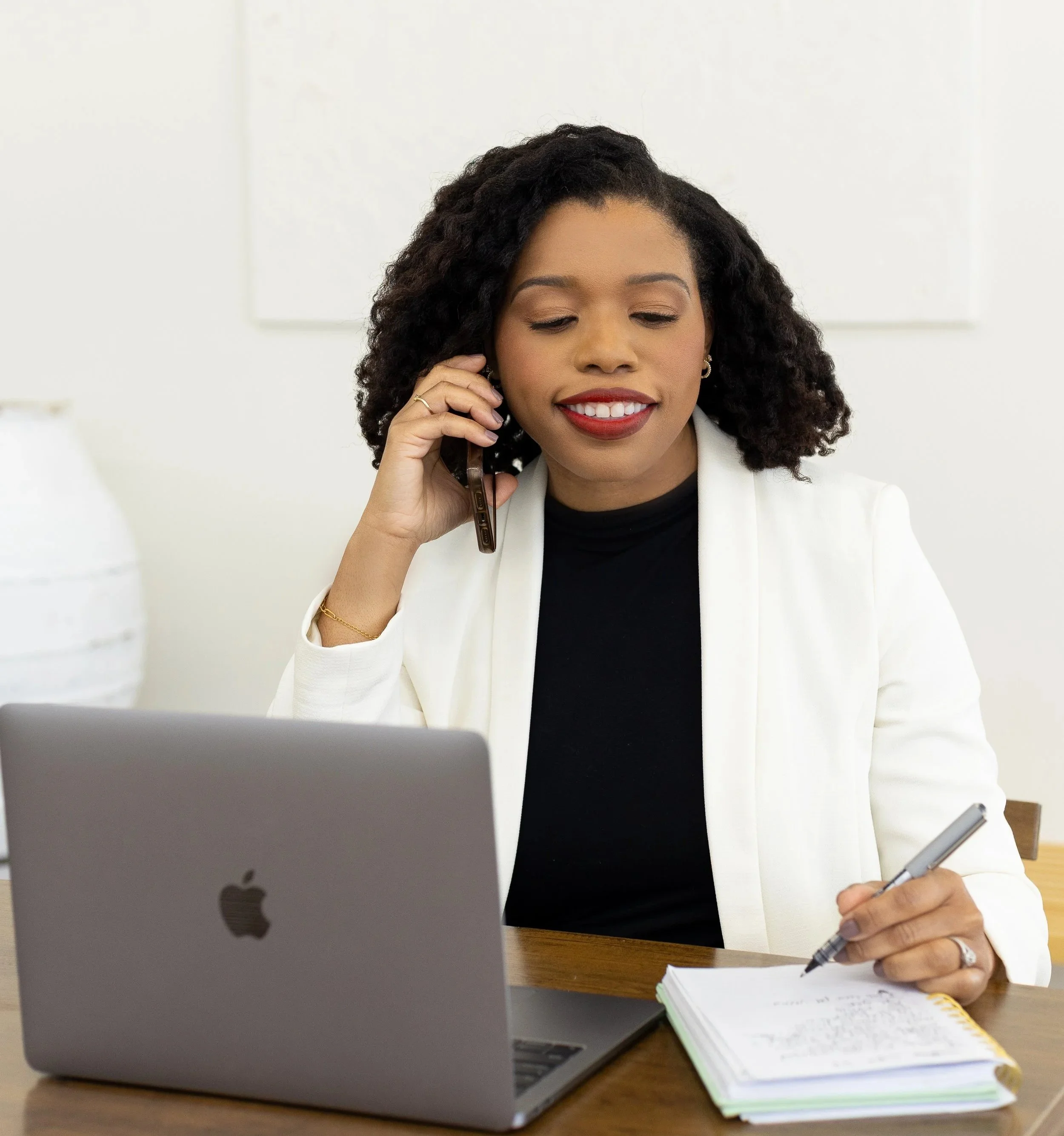 Woman sitting at a desk, talking on a landline phone, writing in a notebook, with an Apple laptop open in front of her.