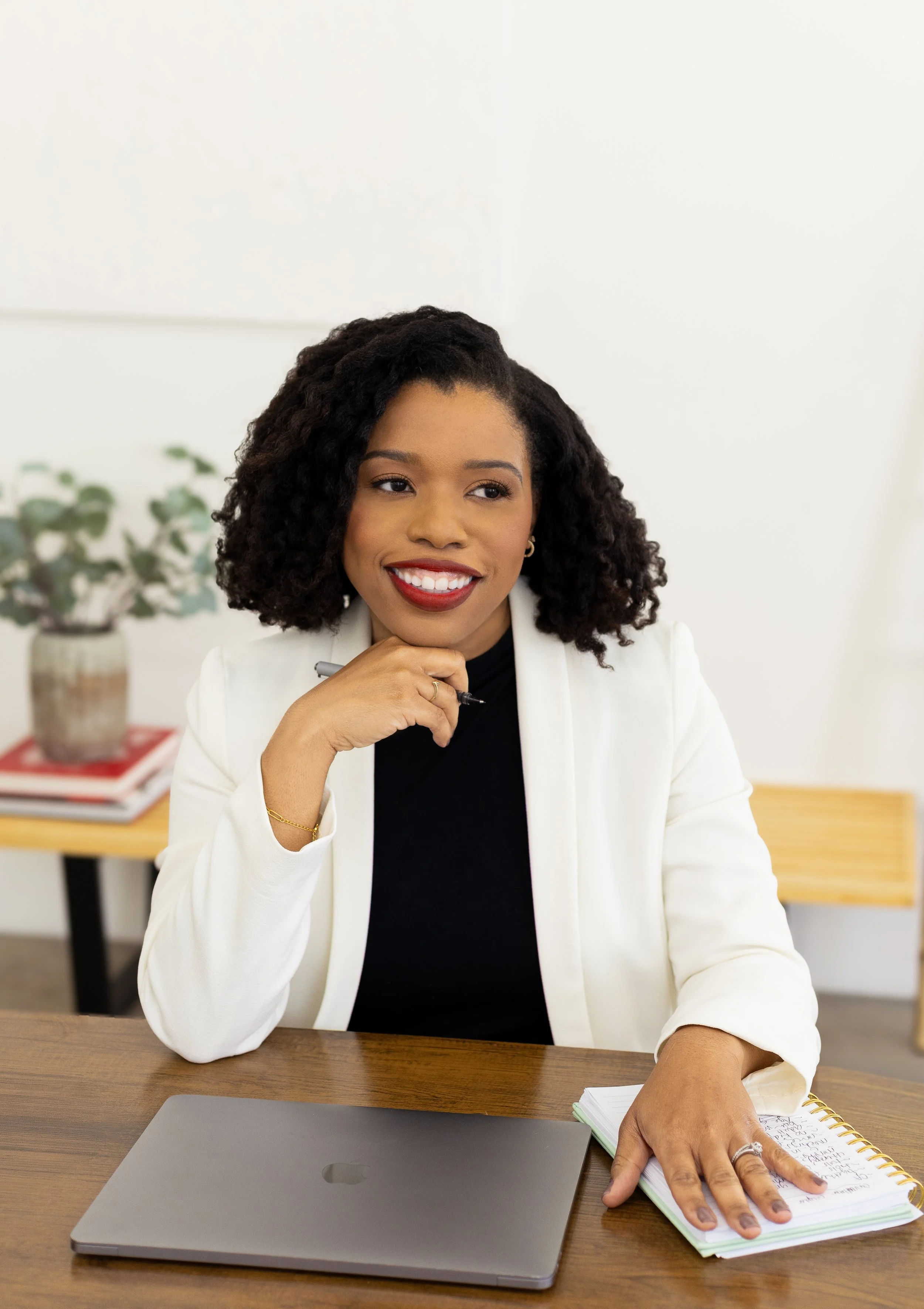 Businesswoman sitting at a desk with a laptop, notebook, and pen, smiling and looking to the side, in an office setting.