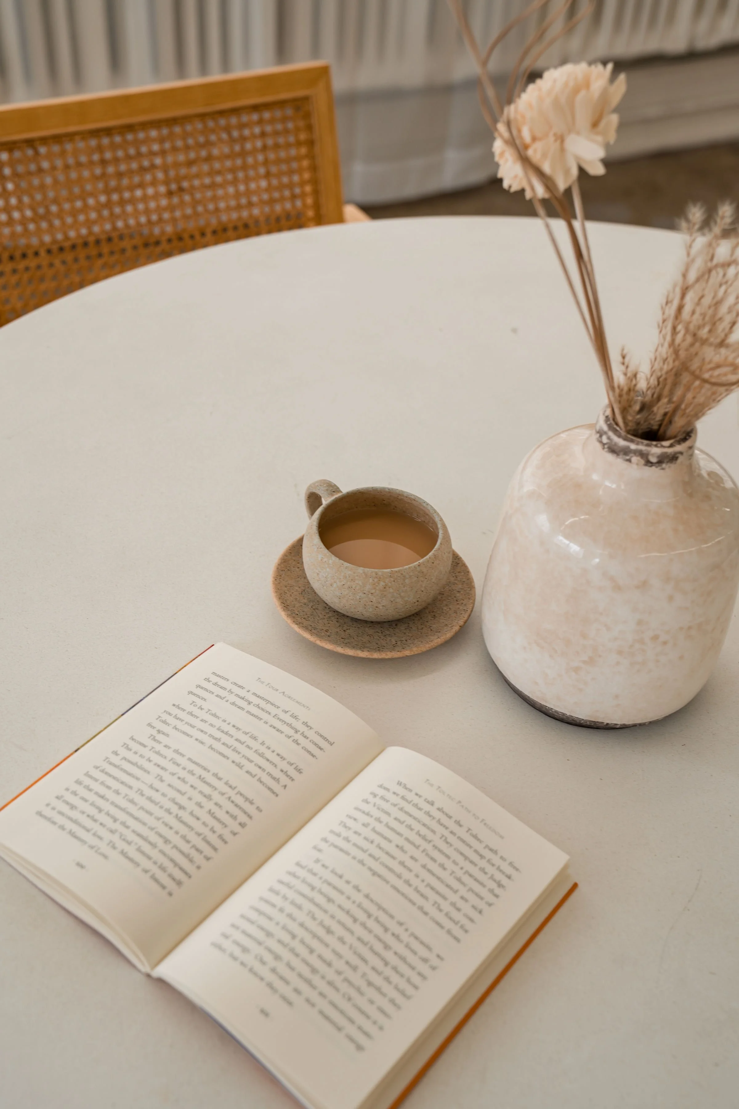 An open book on a white table with a ceramic cup filled with a beverage, a vase with dried flowers, and a partially visible woven chair in the background.