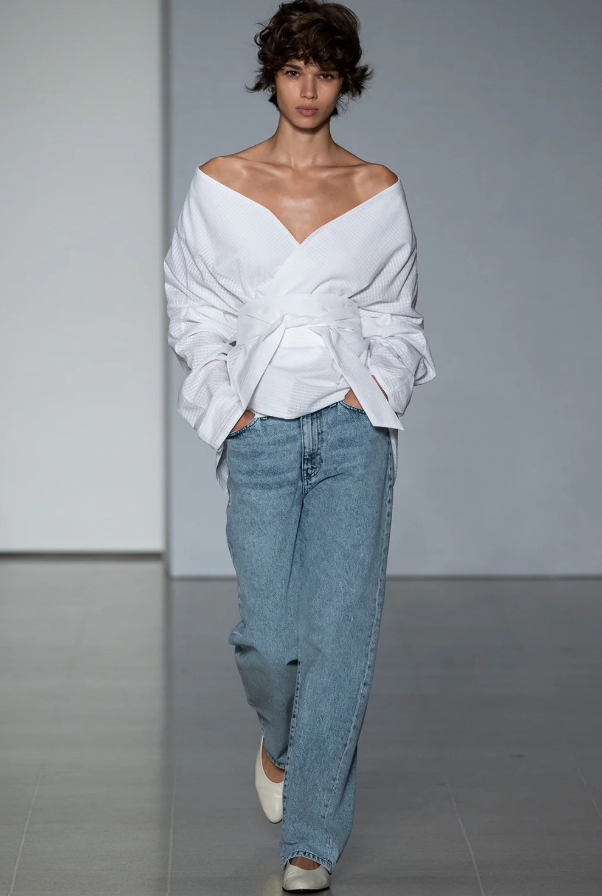 Model wearing white off-the-shoulder wrap top and blue relaxed straight jeans