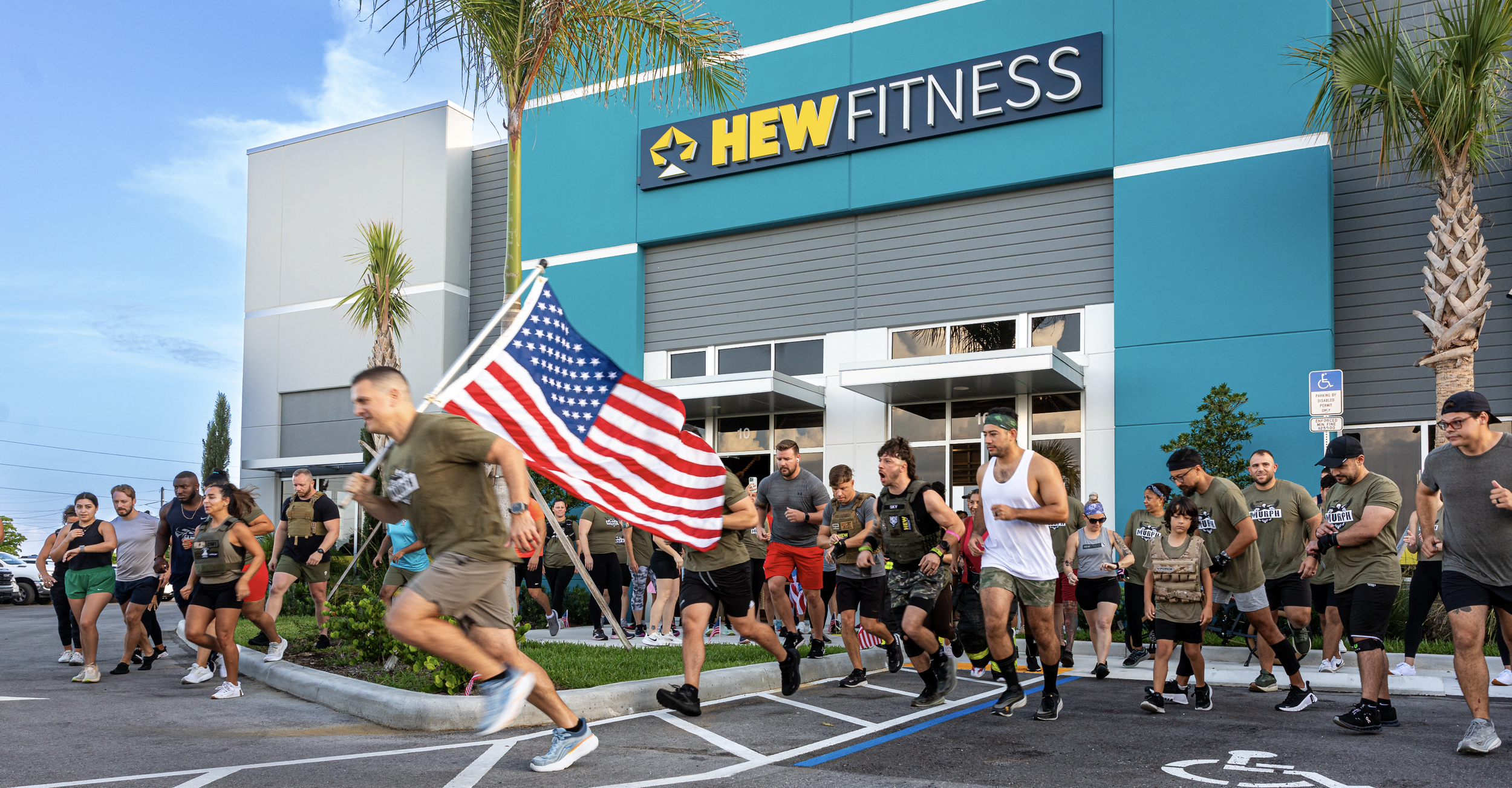 A group of people participating in a running event outside a HEW Fitness gym, with some holding American flags, on a sunny day with blue skies and palm trees.