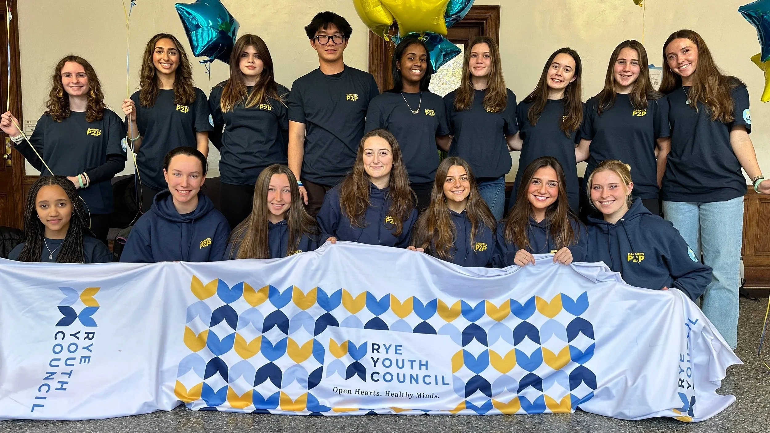 Group of young people gathered together holding a banner that reads 'RYE YOUTH COUNCIL' with the slogan 'Open Hearts. Healthy Minds.' and wearing matching navy and blue clothing, standing indoors with balloons in the background.