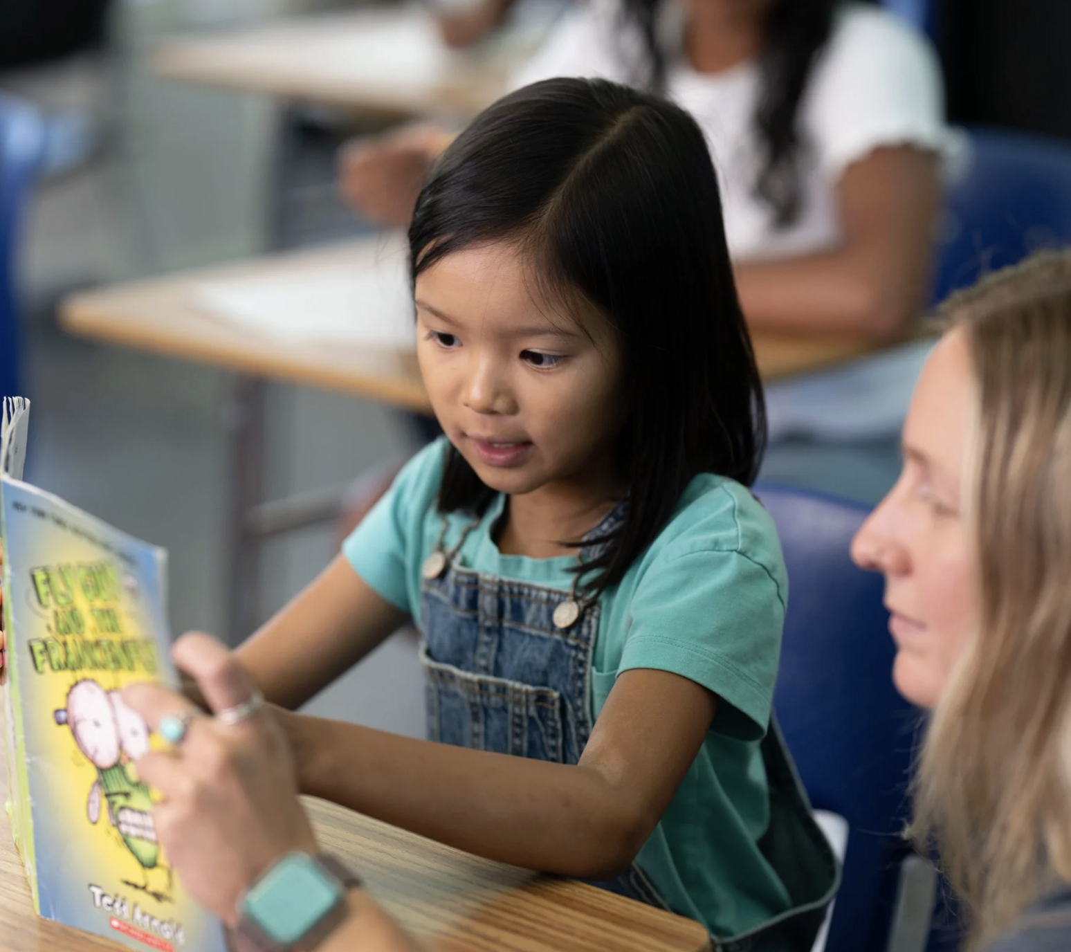 A young girl with straight black hair reading a colorful children's book at a table in a classroom, with an adult and other children in the background.