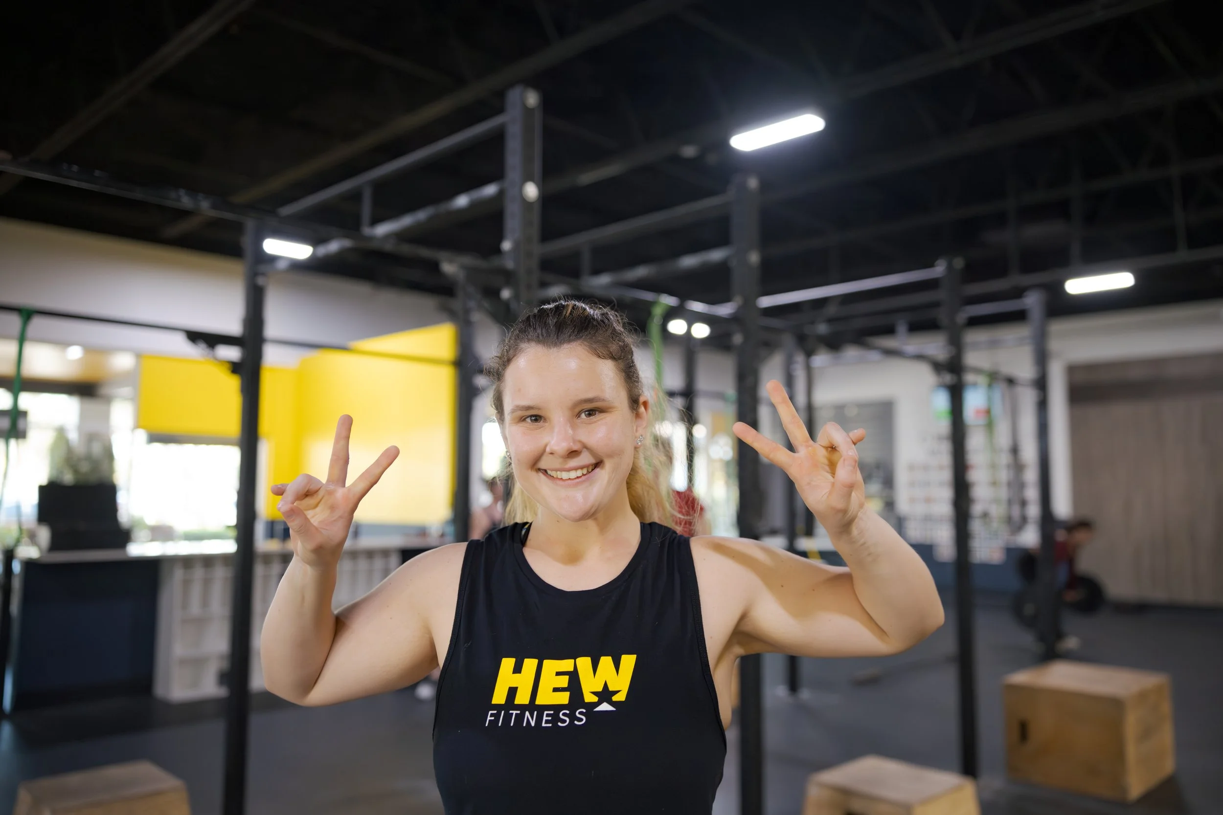 Young woman in workout attire smiling and making peace signs at a fitness gym.