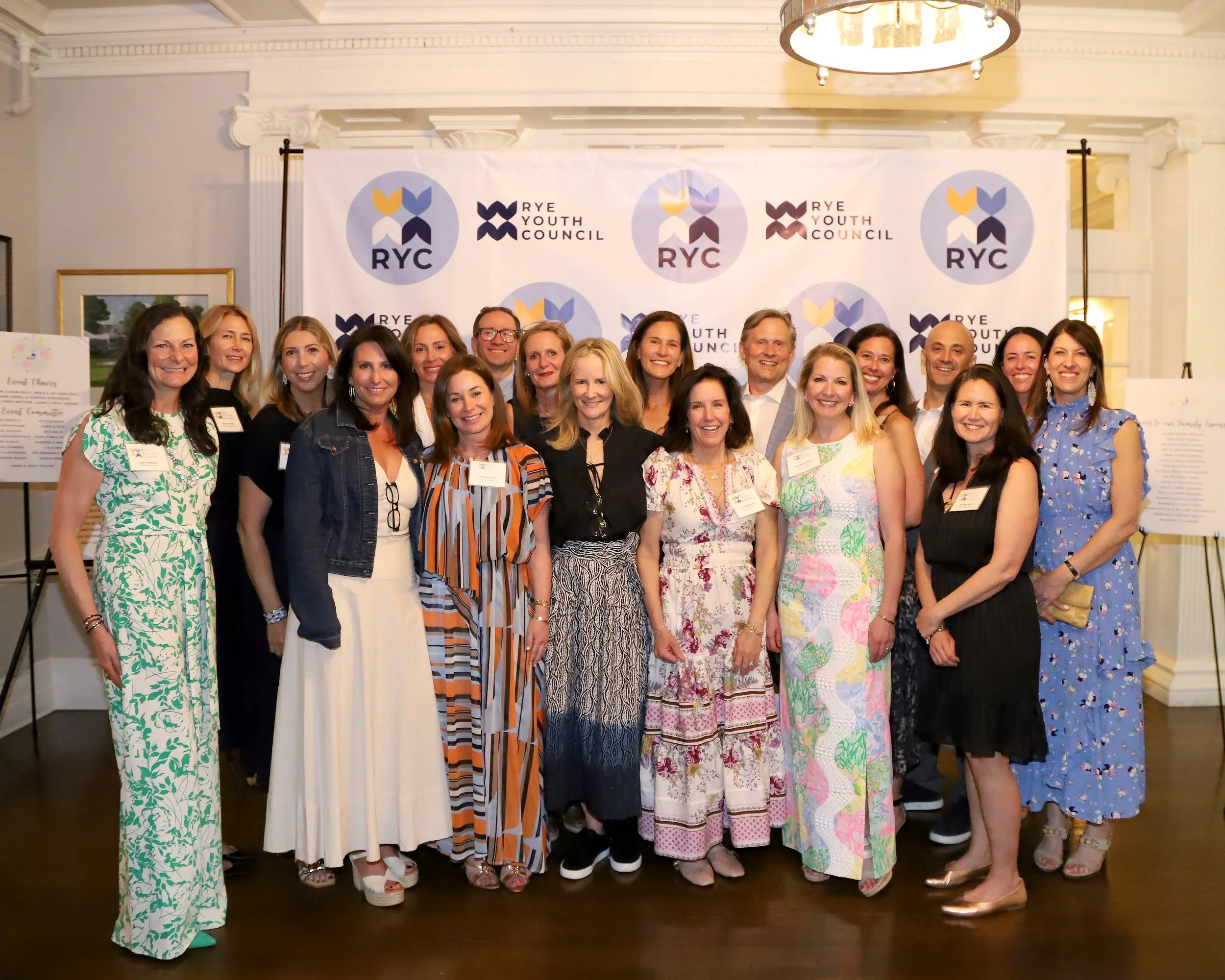 Group of people posing for a photo at a Rye Youth Council event, standing in front of a branded backdrop with the Rye Youth Council logo, in a room with elegant decor and lighting.