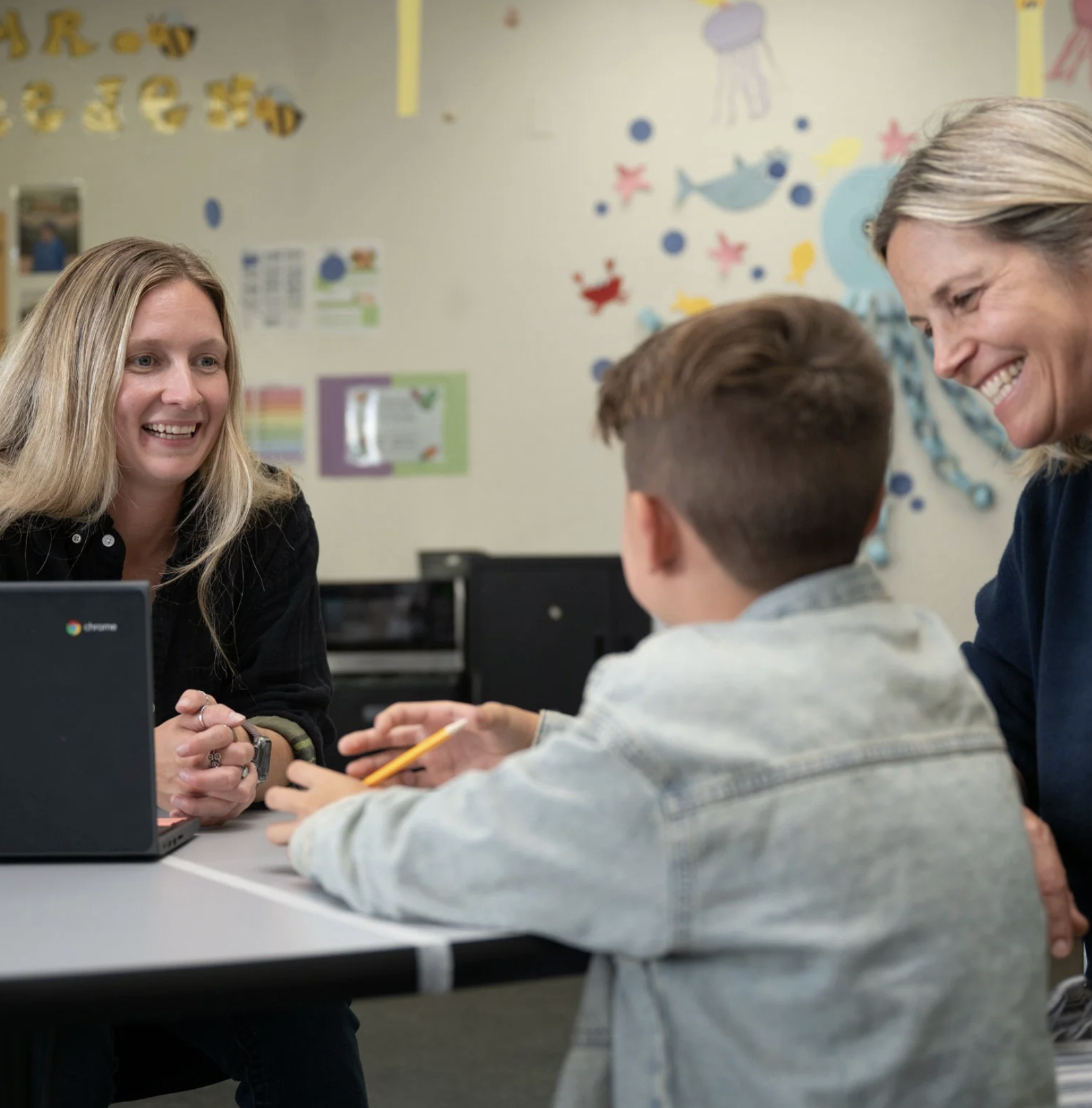 A young boy having a conversation with two smiling women, one with blonde hair and one with gray hair, in a classroom decorated with colorful ocean-themed posters.