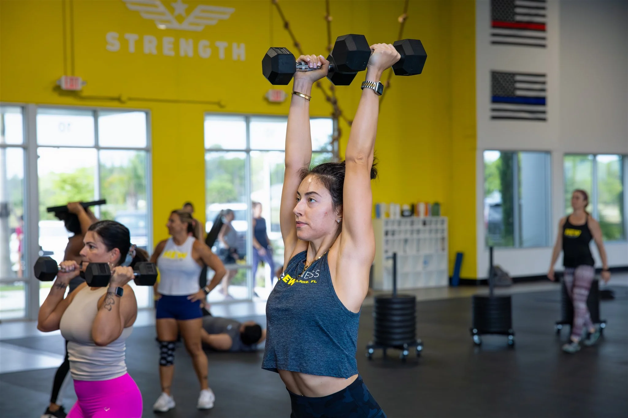 Women participating in a group workout class at a gym, lifting dumbbells overhead, with others exercising in the background.