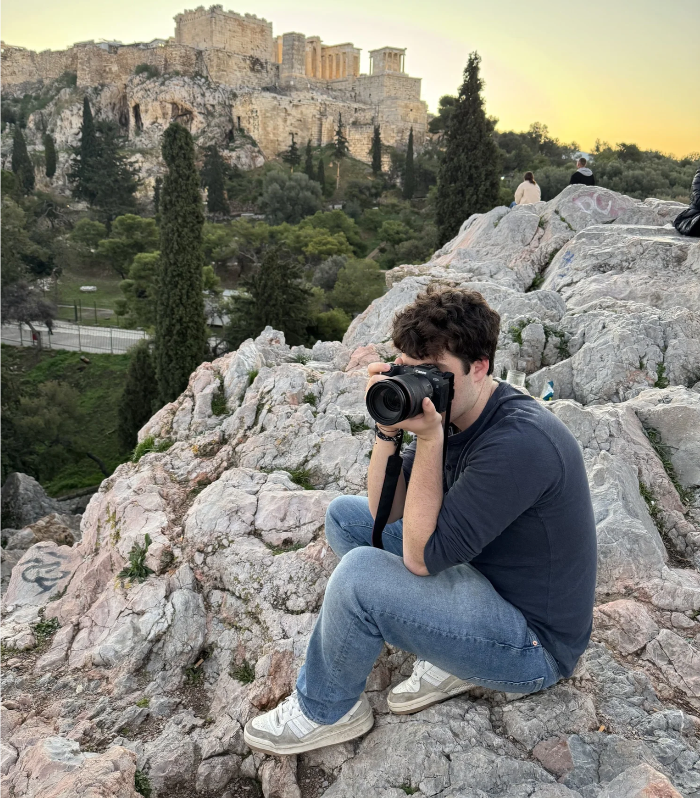 A young man in a dark blue shirt and jeans sits on rocks and takes a photo with a camera. In the background, the Acropolis of Athens is visible at sunset, with ancient ruins and tall trees.