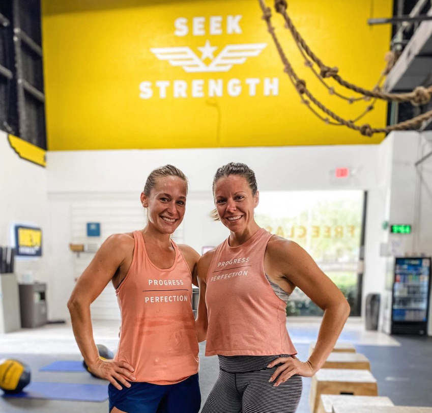 Two women smiling inside a gym, wearing workout tank tops that say 'Progress' and 'Perfection.' Gym equipment and a large yellow wall with the words 'Seek Strength' are visible in the background.