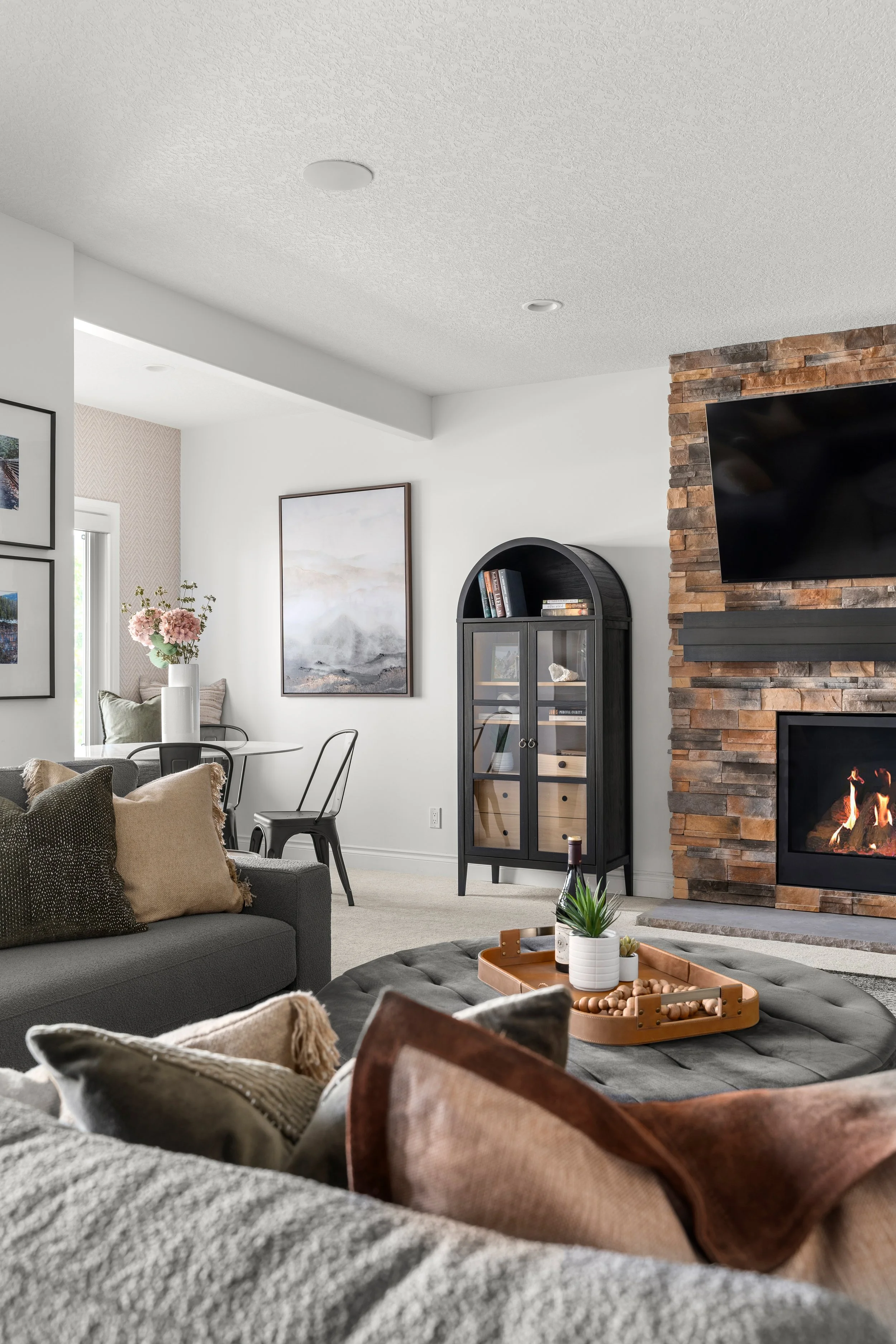 A cozy living room with a stone fireplace, a mounted flat-screen TV, a black display cabinet, a gray sofa with pillows, a round tufted ottoman with a tray, and a dining area with a table, chairs, and a vase of pink flowers.