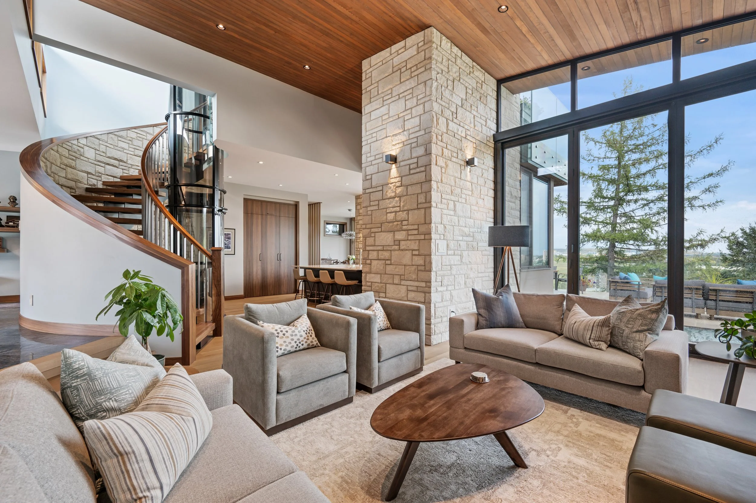 Living room with a large stone fireplace, beige sofas and armchairs, a wooden coffee table, large glass windows showing outdoor trees, a wooden ceiling, and a staircase with a glass elevator.