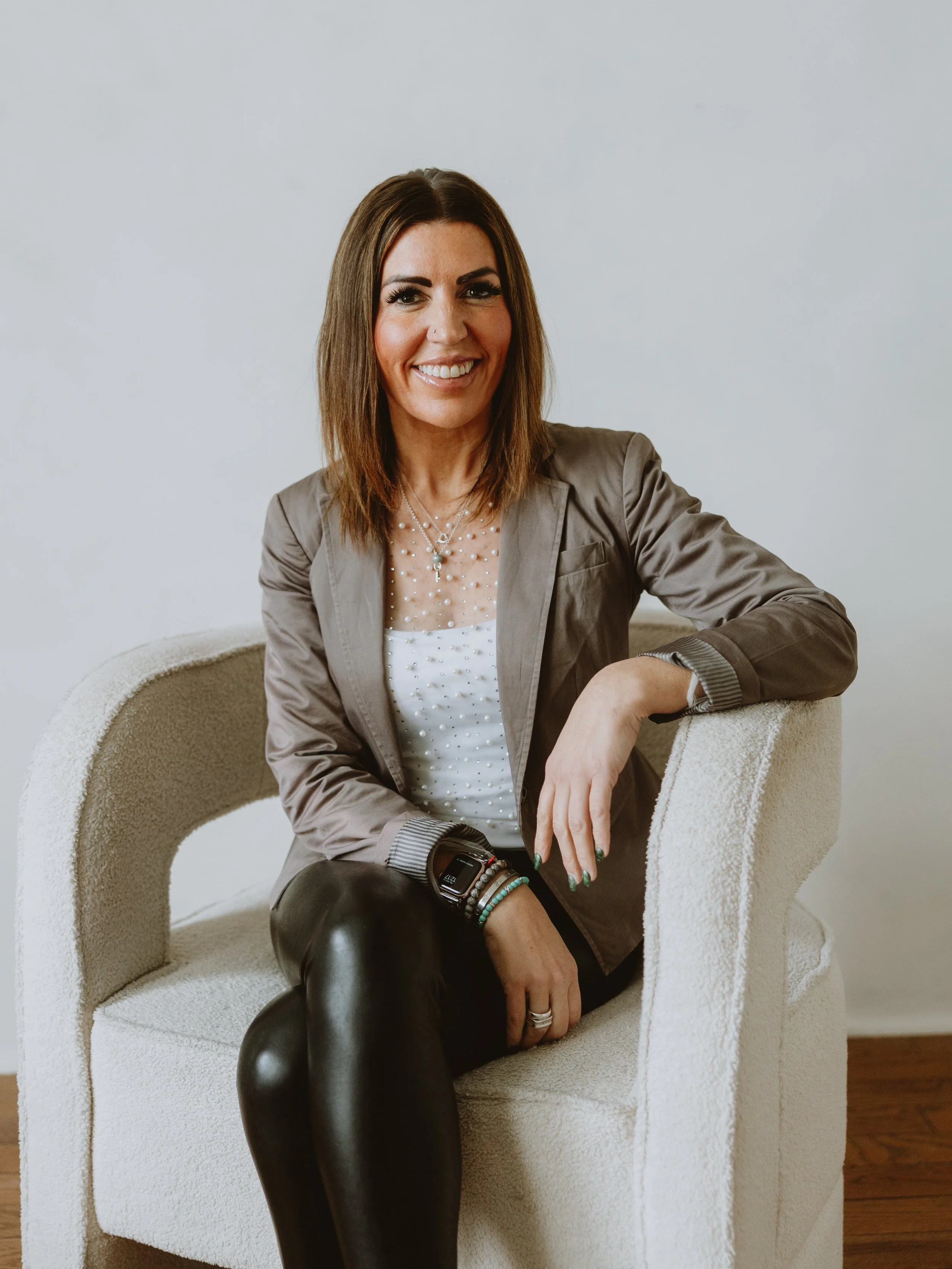 A smiling woman with shoulder-length brown hair sitting on a white textured armchair with a plain white wall in the background.