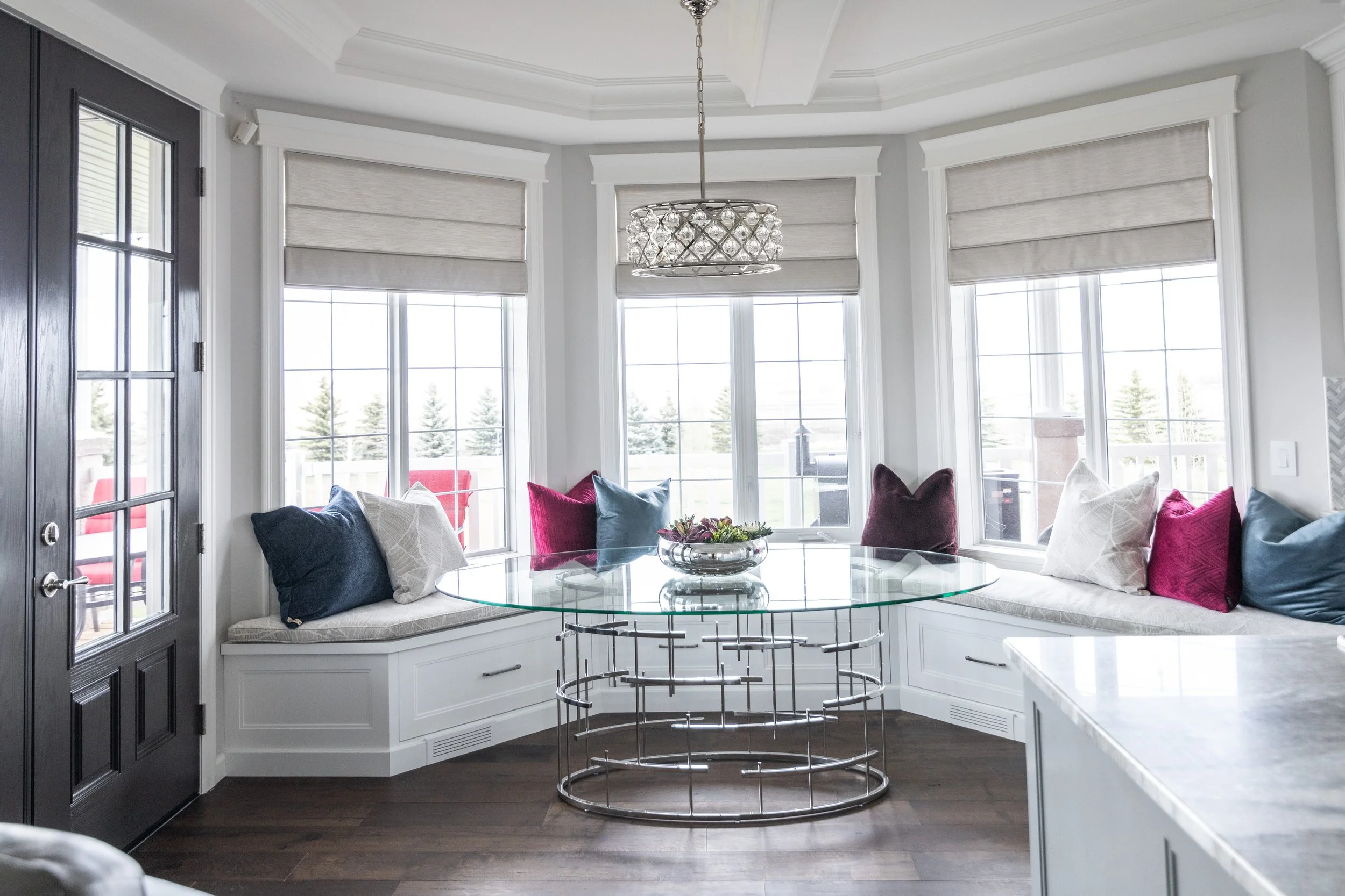 Bright sunroom with bay windows decorated with plush pillows and a glass-top table on dark wooden flooring.