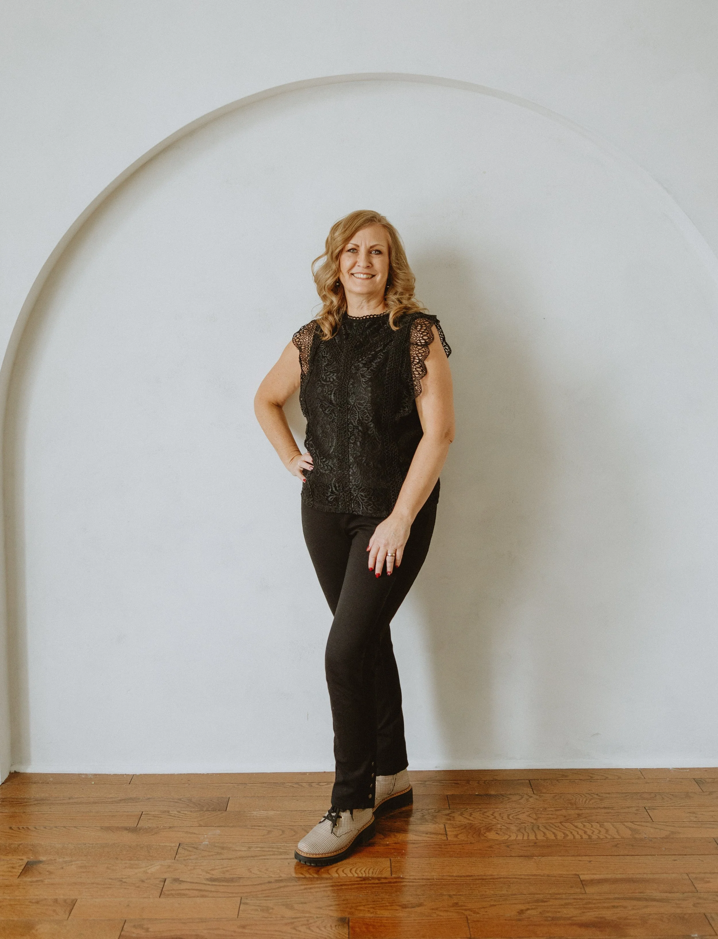 A woman with wavy blonde hair posing in front of a white wall with an arch. She is wearing a black lace top, black pants, and beige shoes, smiling with her hand on her hip.
