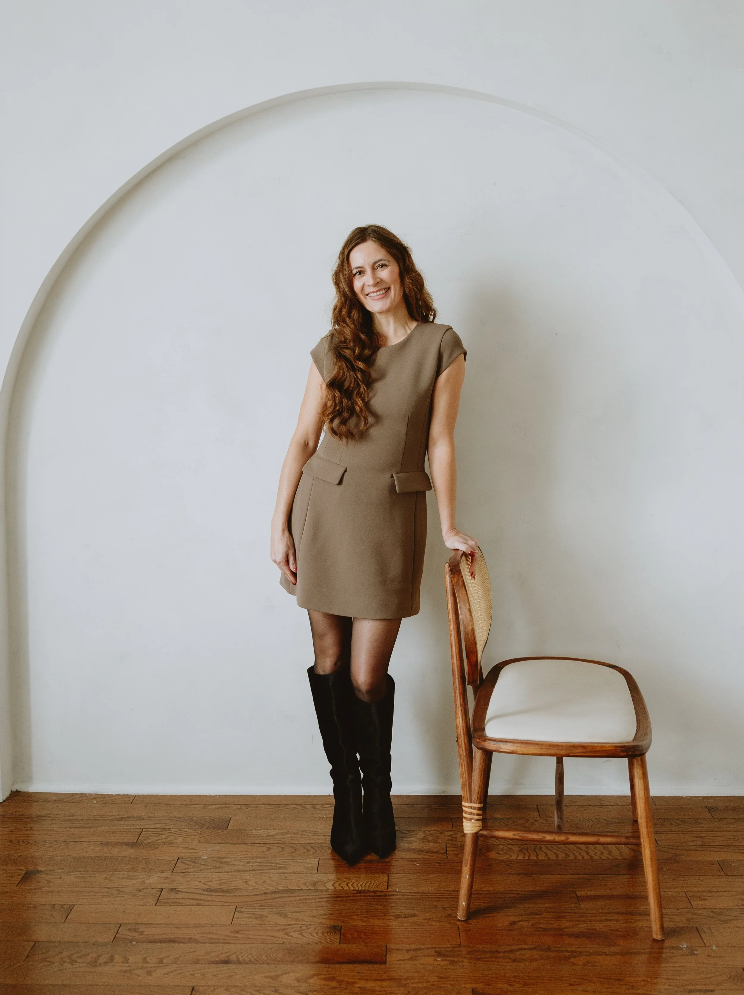 A woman with long wavy brown hair smiling, wearing a taupe dress, black knee-high boots, standing next to a wooden chair with a white cushion, in a room with hardwood floors and a white wall with an arch design.