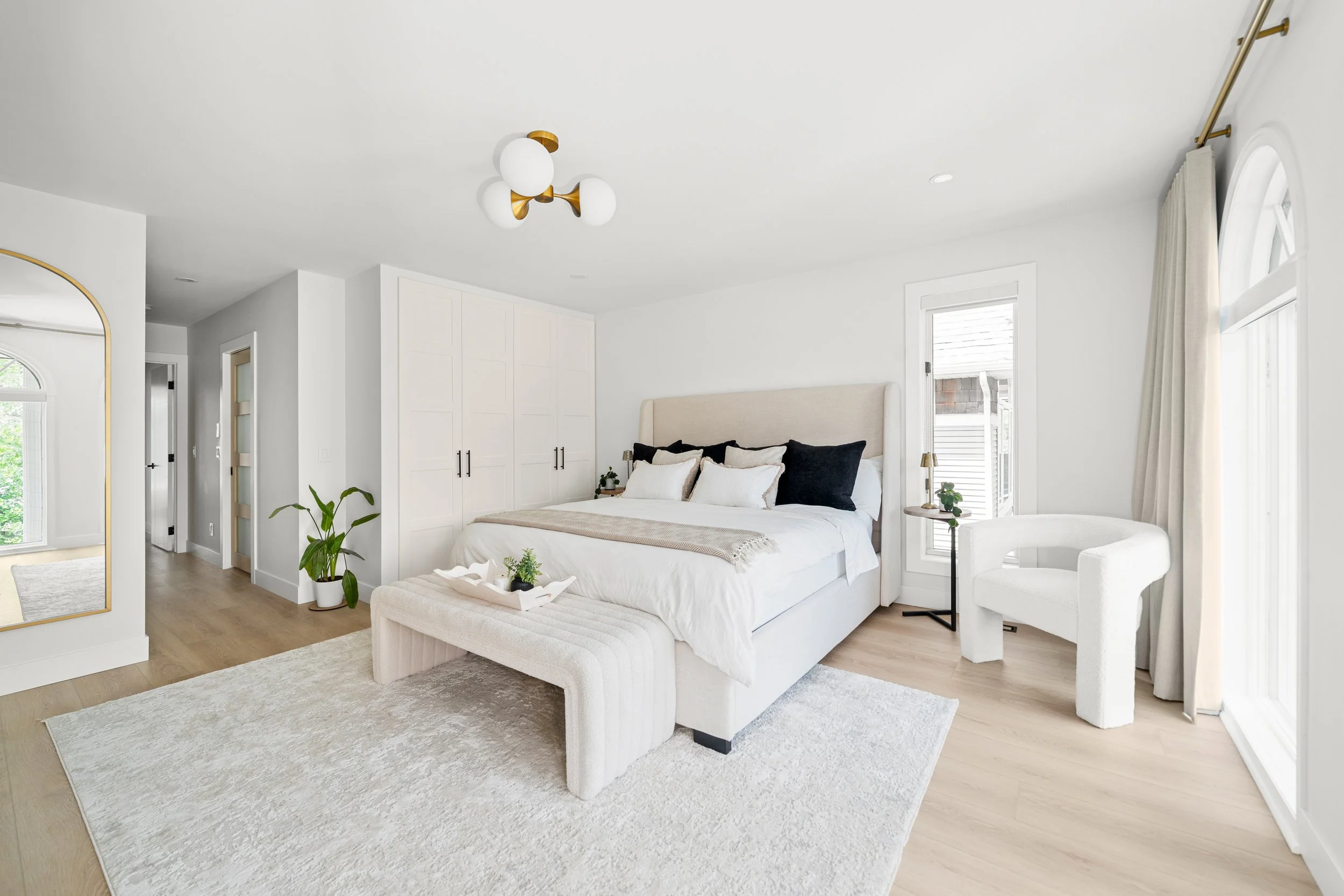 Bright modern bedroom with white walls, a large bed with white and black pillows, a beige upholstered headboard, a white bench at the foot of the bed, and a white armchair near windows with white curtains. There are green plants, a round side table, and a gold and white ceiling light.