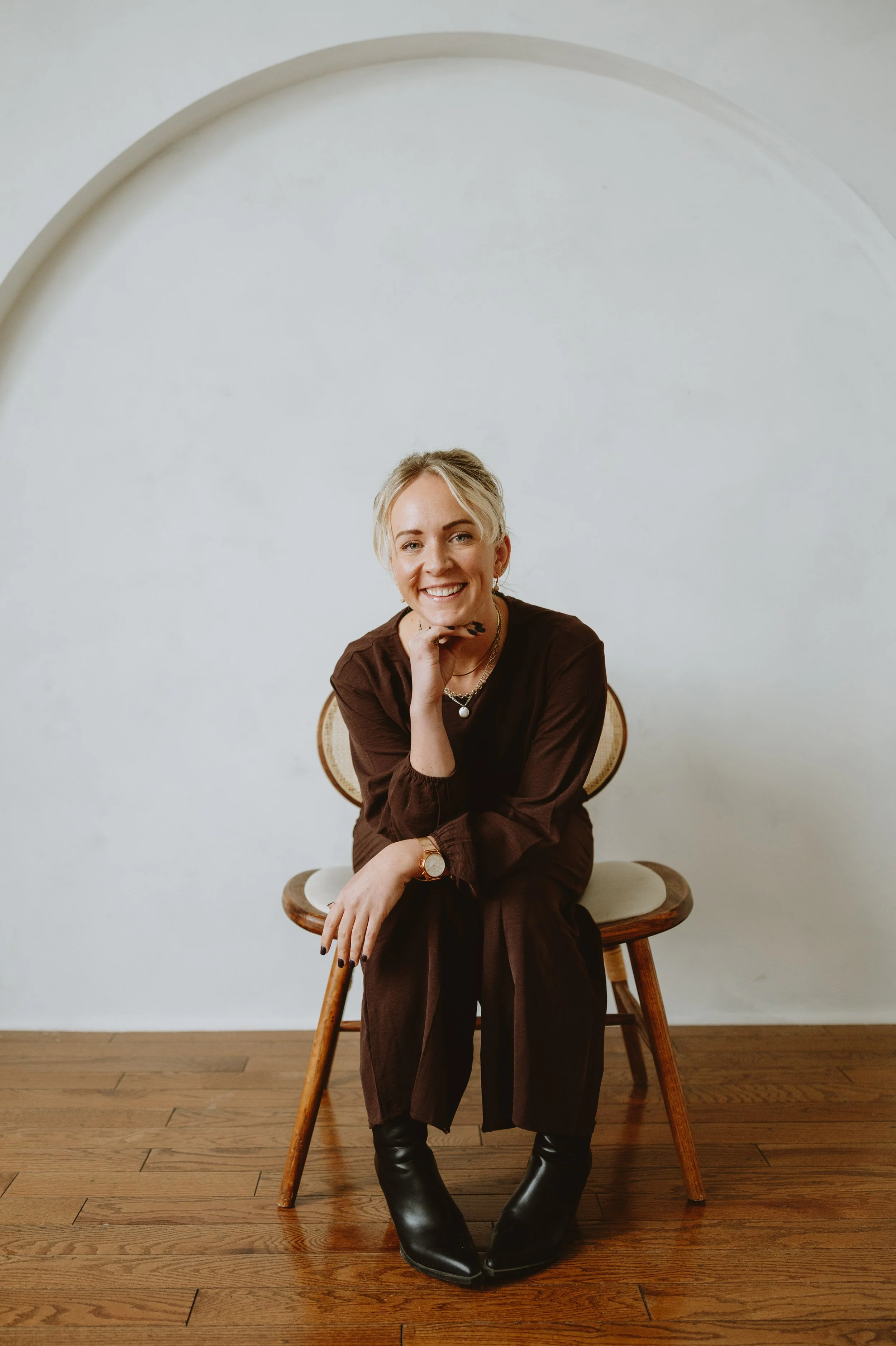 A woman with blonde hair and a brown outfit sitting on a wooden chair, smiling at the camera.