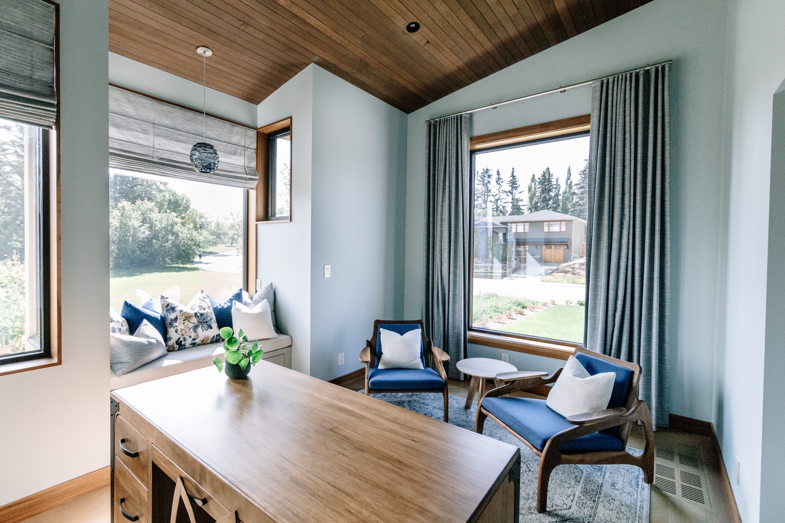 Modern living room featuring large windows with curtains, blue chairs with pillows, a built-in window seat with pillows, a wooden table, and outdoor views of a suburban neighborhood.
