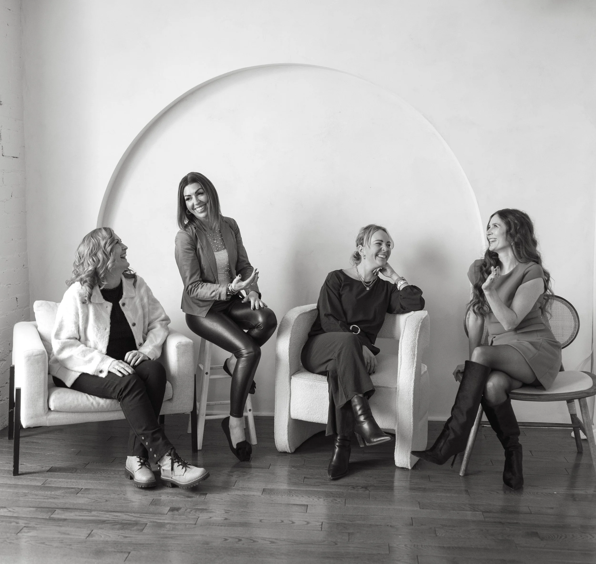 Four women sitting and standing on chairs in a room, engaging in conversation and laughter. Black and white photo.