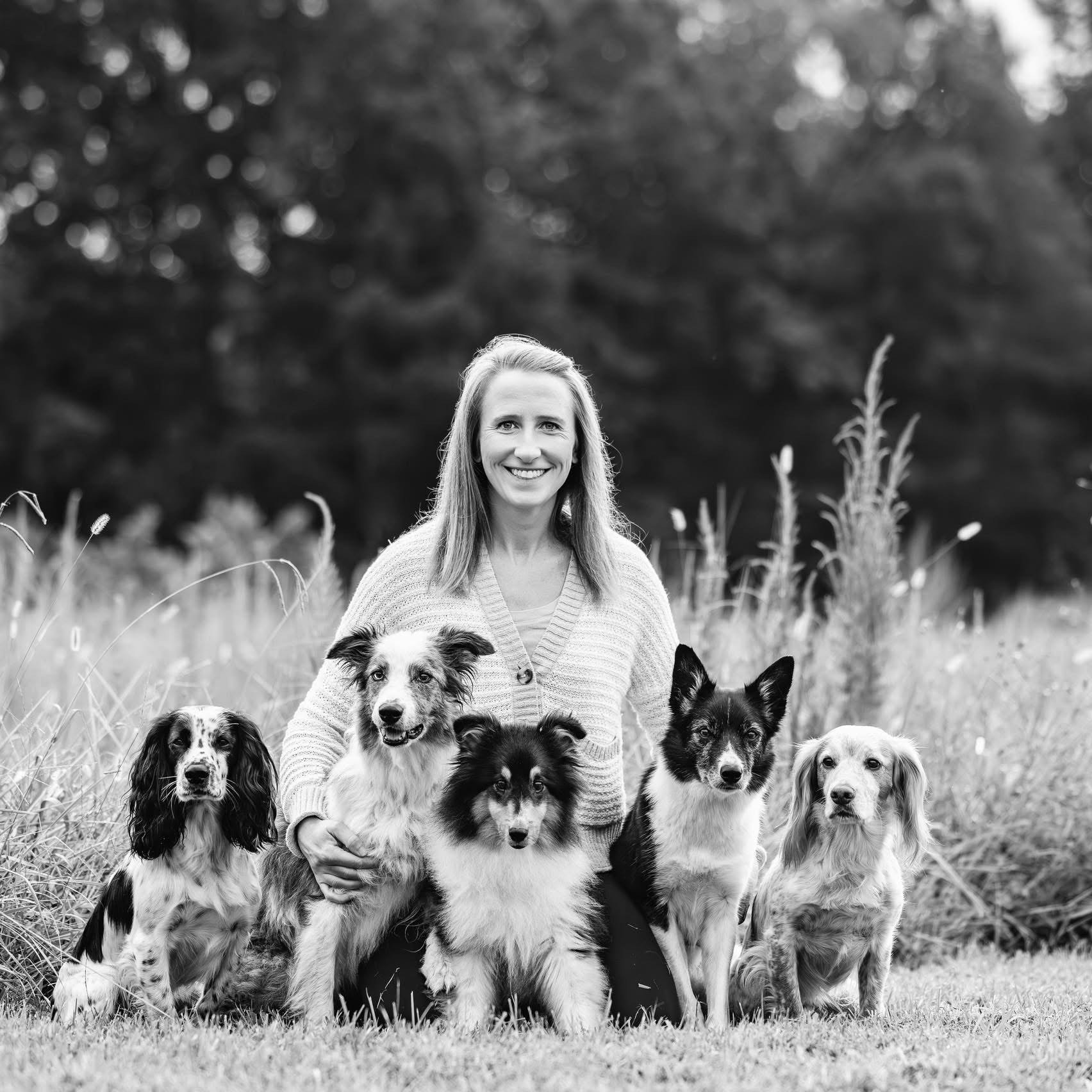 A woman smiling, kneeling outdoors in a grassy field with five dogs of various breeds, all looking toward the camera, black and white photo.