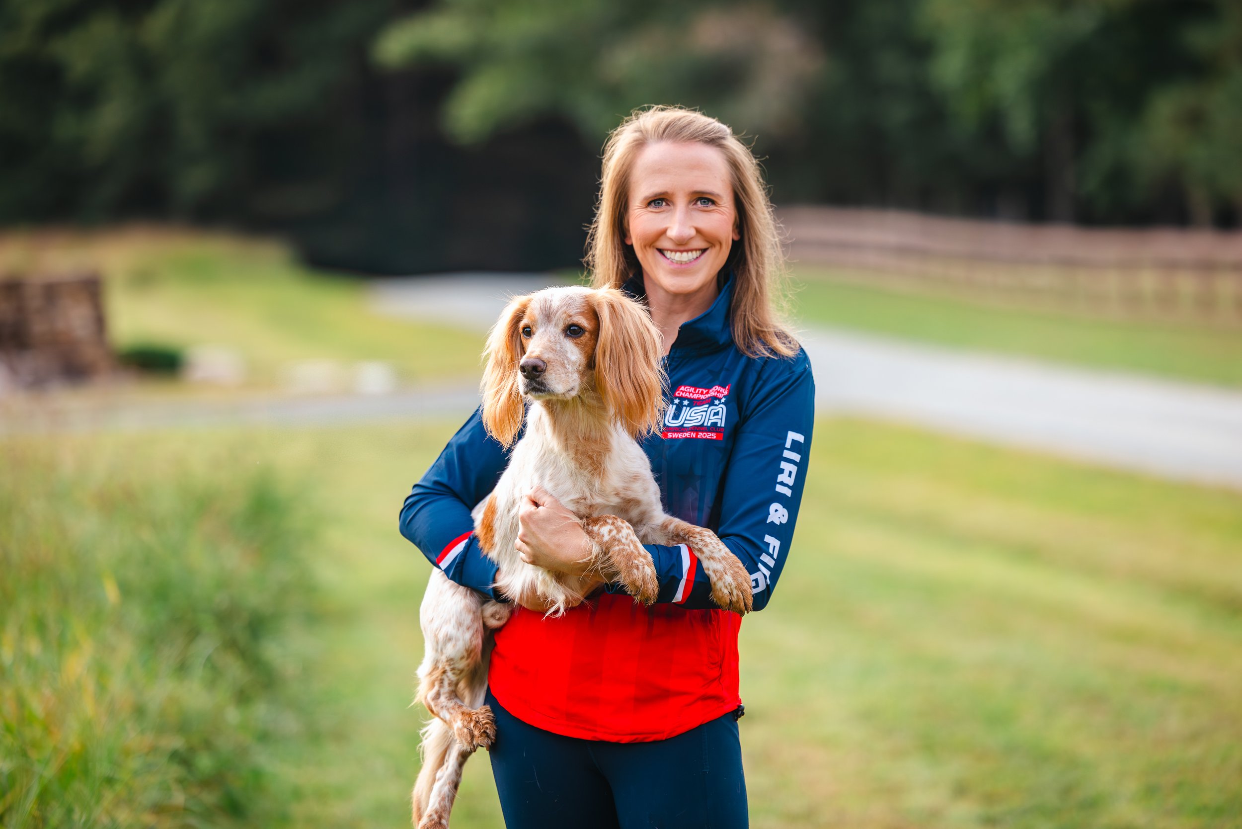 A woman with long blonde hair smiling and holding a small dog outdoors on a grassy trail with trees in the background.