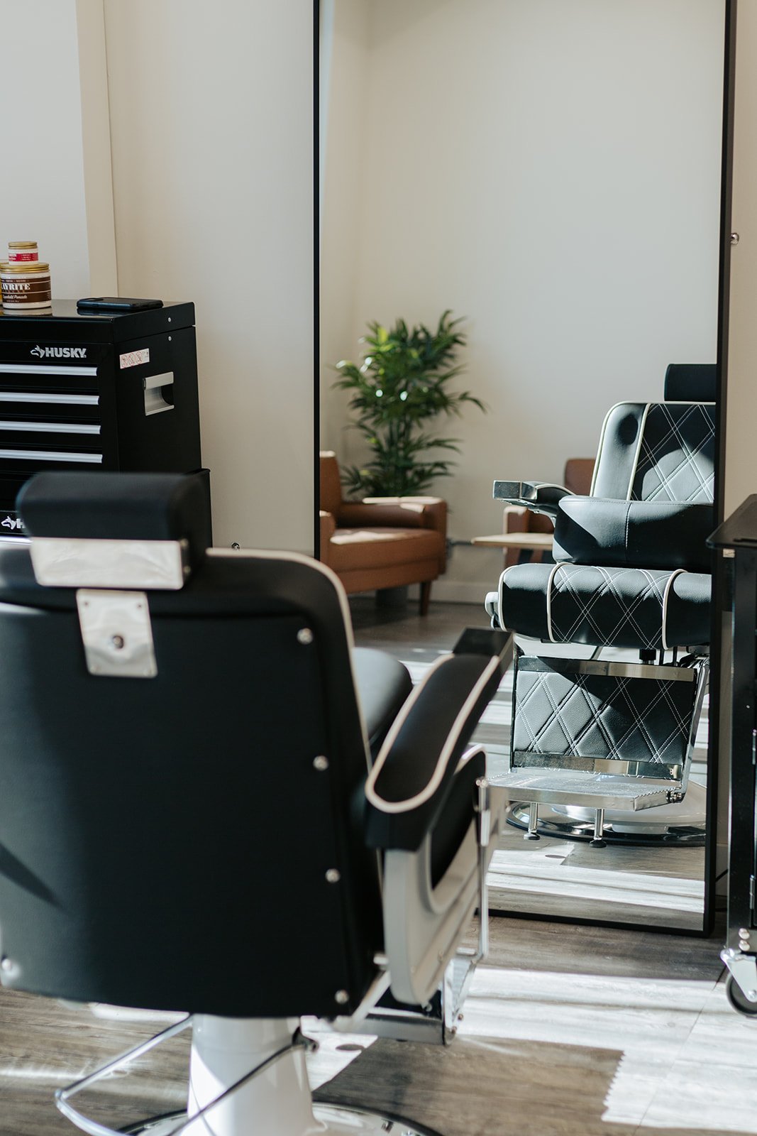 Photo of a black leather barber chair with white stitching and chrome hardware, reflected in a floor length mirror. A seating area can be seen in its reflection.