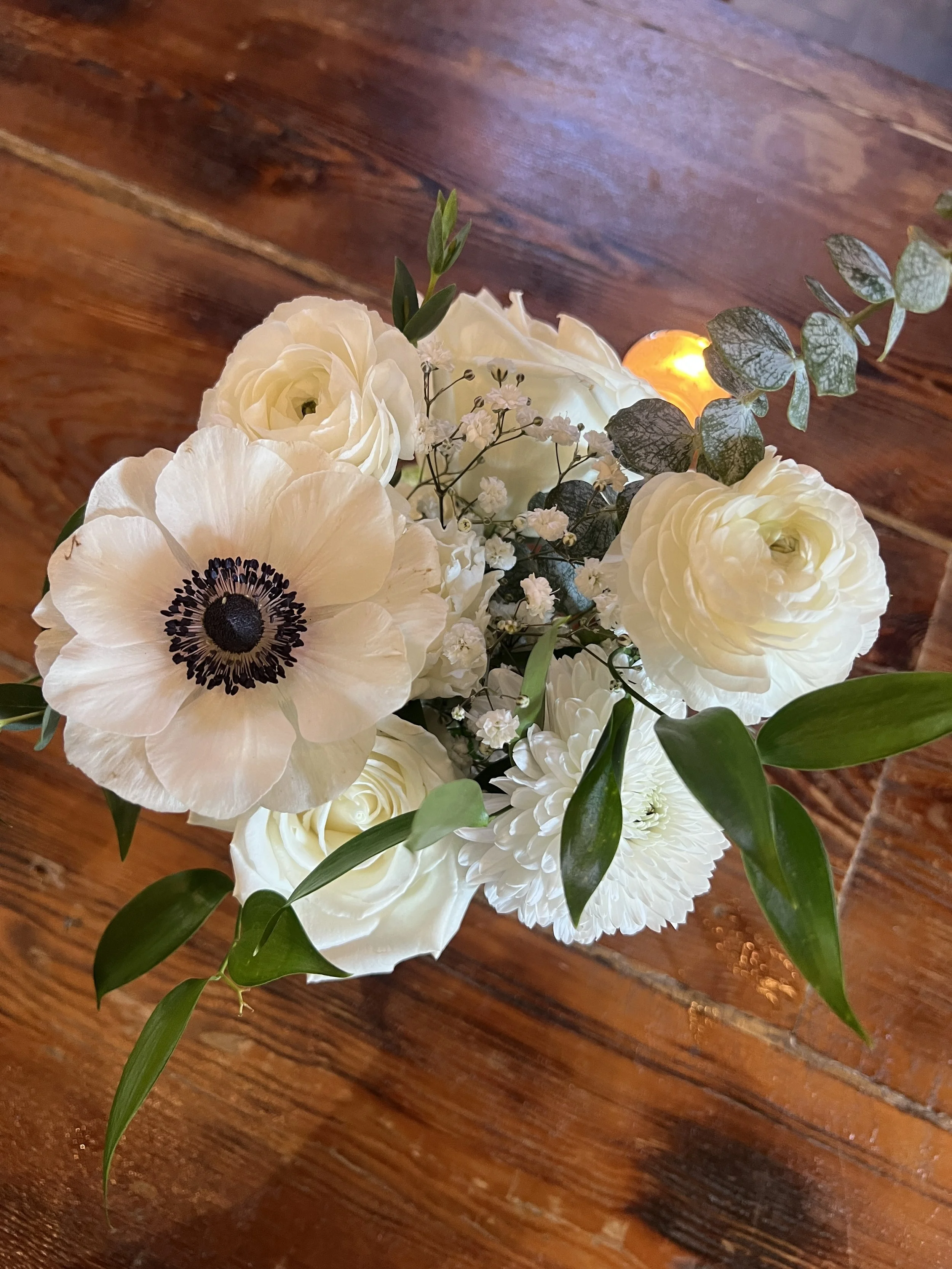 A baby shower decorative floral arrangement of white focal flowers including anemones, roses, and baby's breath, with green leaves, on a wooden bar top in Chester County, PA