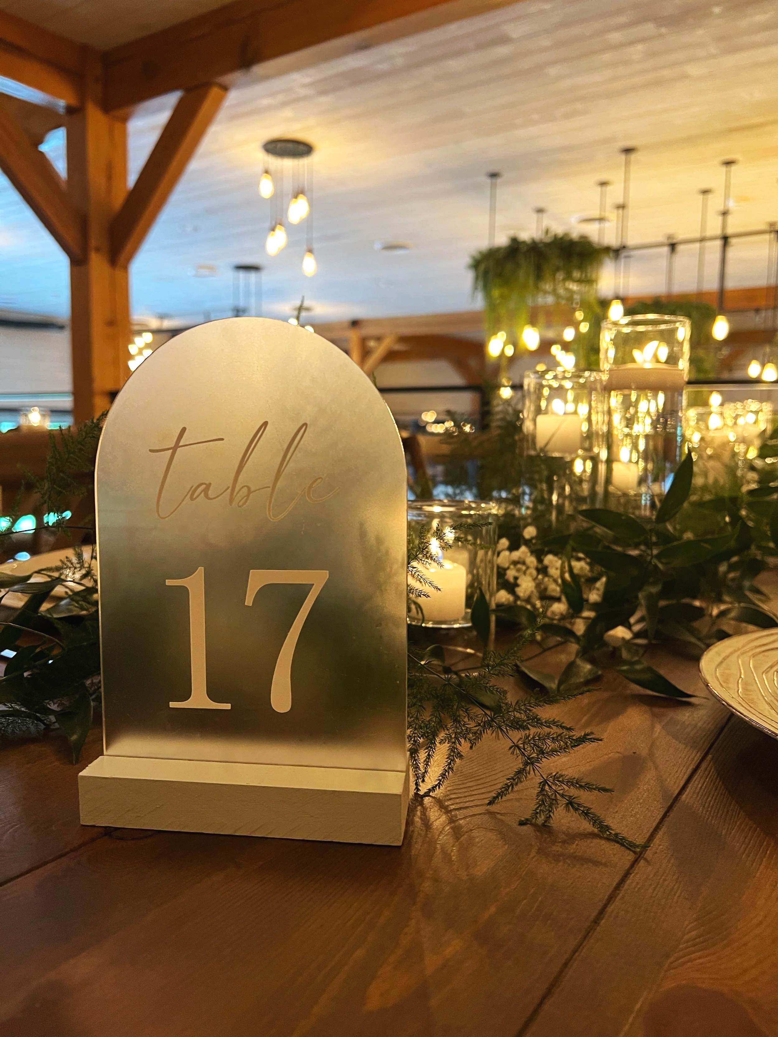 Frosted arch acrylic wedding table number sign created by Fern Hill Design displaying “Table 17” with elegant script lettering, styled on a wooden reception table with greenery and candlelight at a romantic wedding reception in western PA