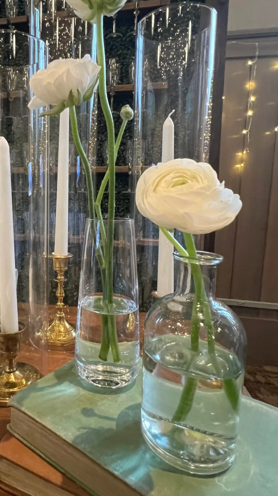 White ranunculus flowers in small rental glass bud vases placed on vintage books, surrounded by taper candles in brass candle holders in clear hurricane sleeves on a rustic wooden table with champagne wall in the background setup in Chester County PA