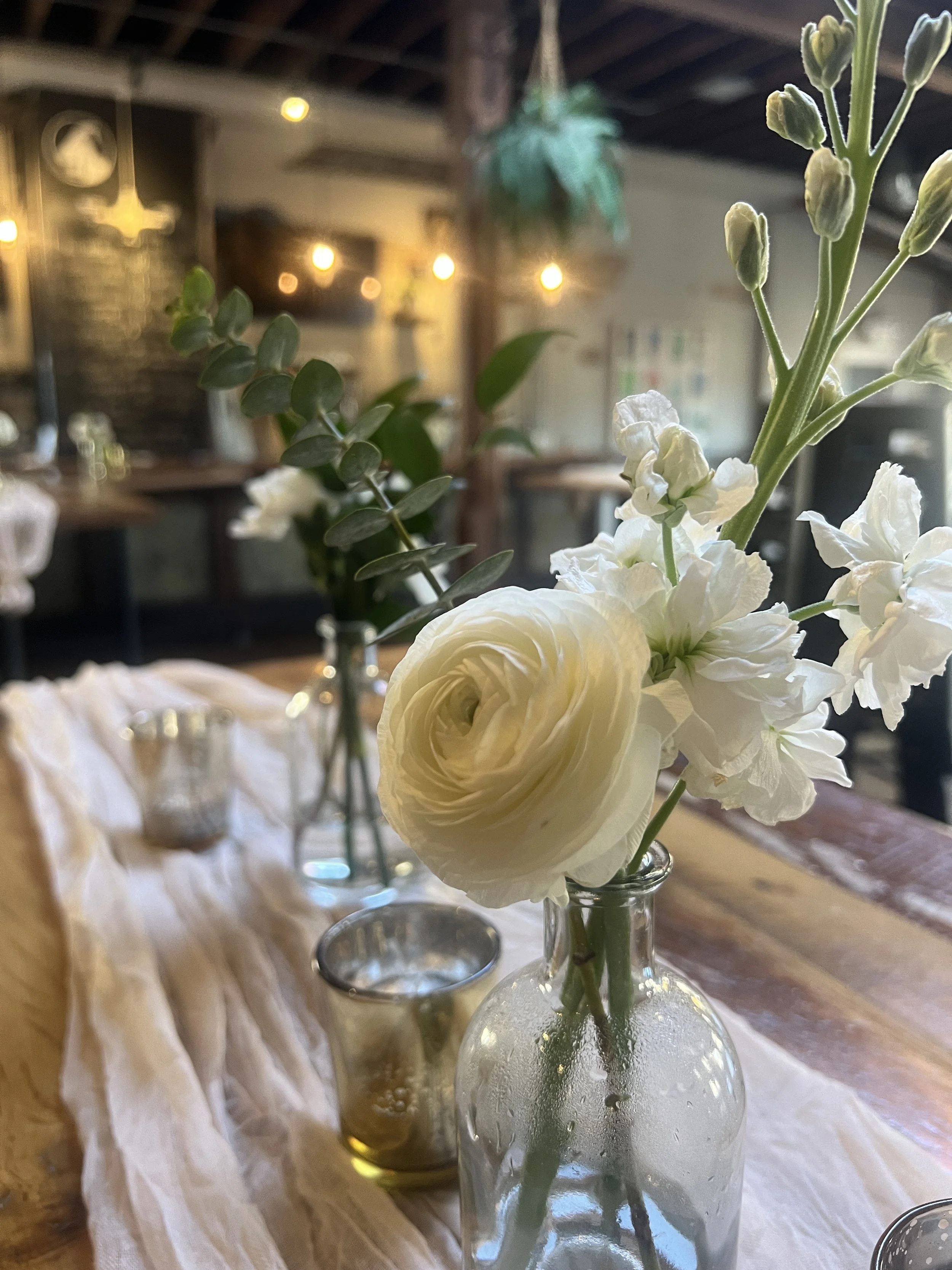 Luxury bud vase flower arrangement with ranunculus, stock and eucalyptus in a small glass bud vase, alongside mercury glass votive candles and setup on a blush colored cheesecloth table runner and rustic wooden tabletop