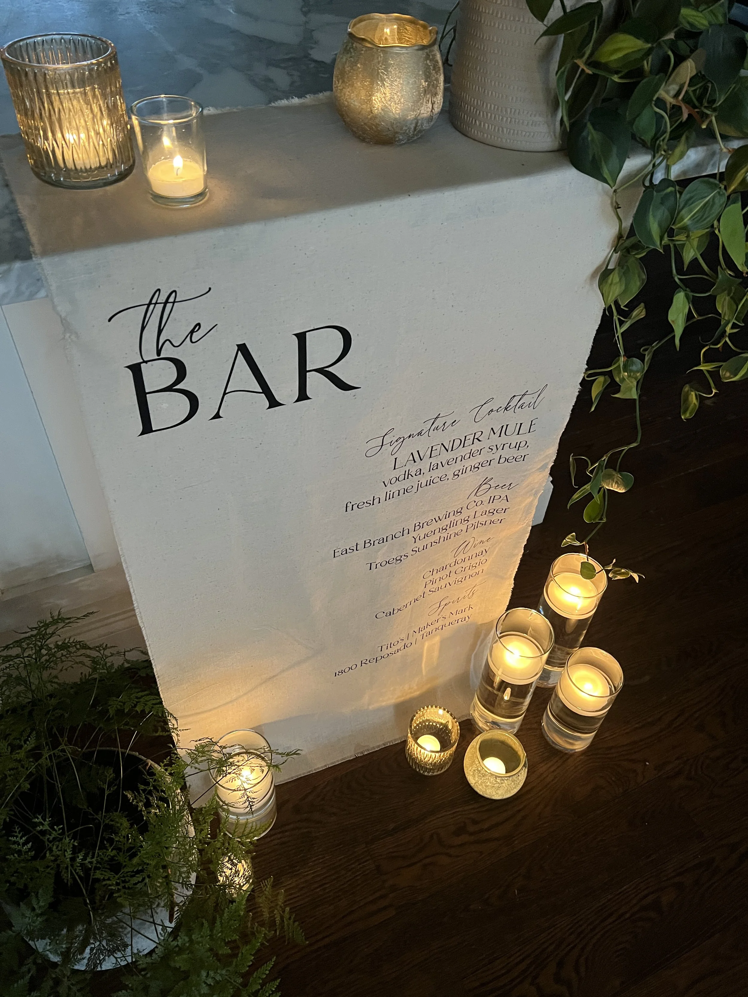 Custom linen bar sign made of light canvas fabric featuring modern script font, with signature cocktail, beer, wine and spirits surrounded by candles and natural plant decor in Chester County PA