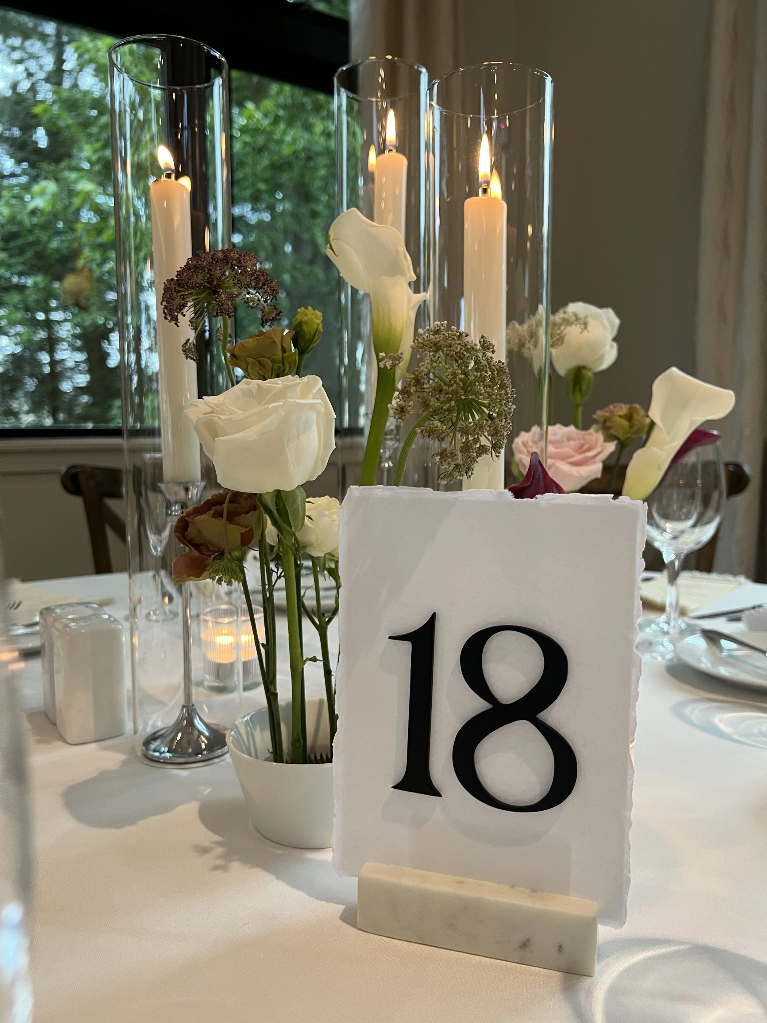 Wedding table numbers made of raised wooden numbers, watercolor paper with deckled edge, and marble number holder, taper candles with hurricanes in background and modern and romantic luxury wedding florals