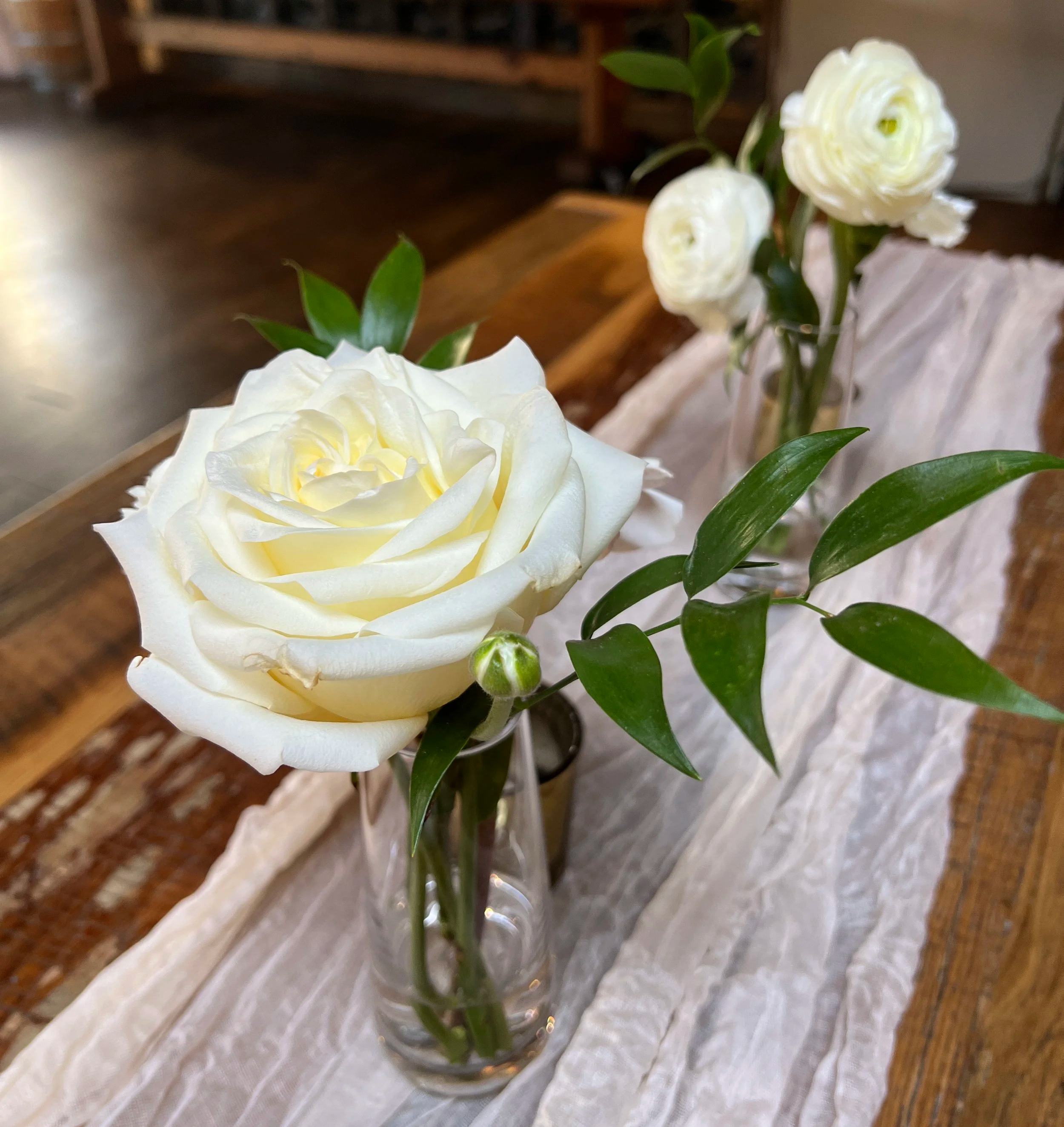Three white roses in small clear glass vases arranged on a rustic wooden table with a white table runner at Main Line Baby Shower, Chester County, PA.
