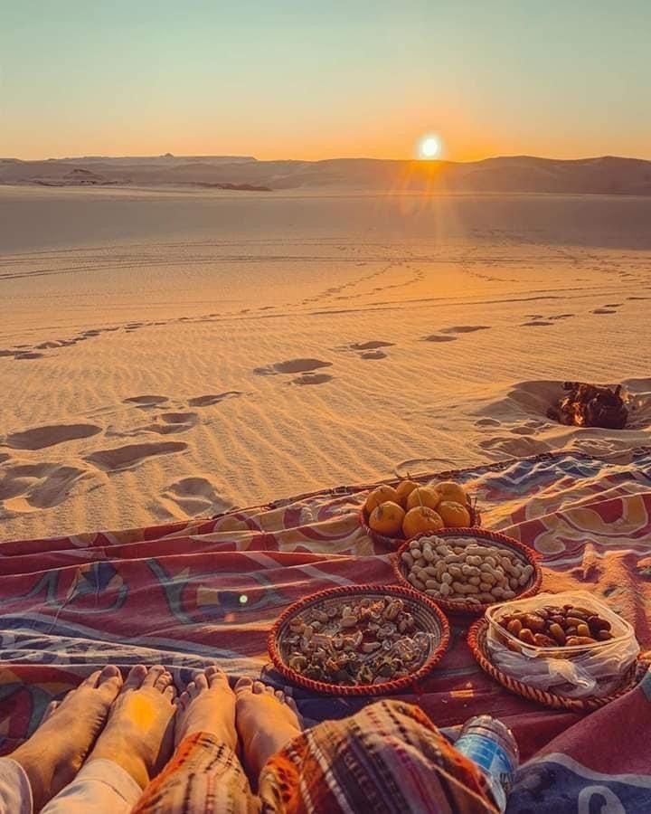 Sunset over a desert with a blanket and various bowls of snacks in the foreground, and footprints in the sand.