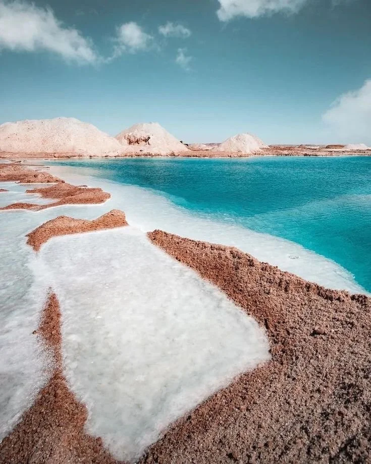 Salt flats with white salt deposits, brownish-red terrain, and a large body of blue water under a partly cloudy sky.