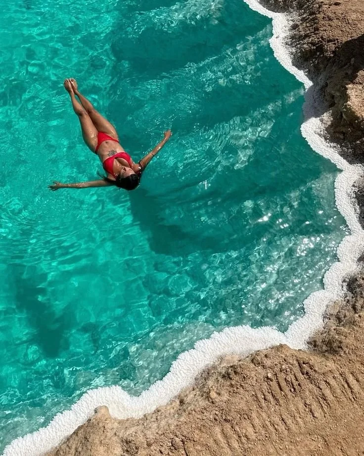 A woman in a red bikini is floating on her back in a turquoise swimming pool near rocky edges.