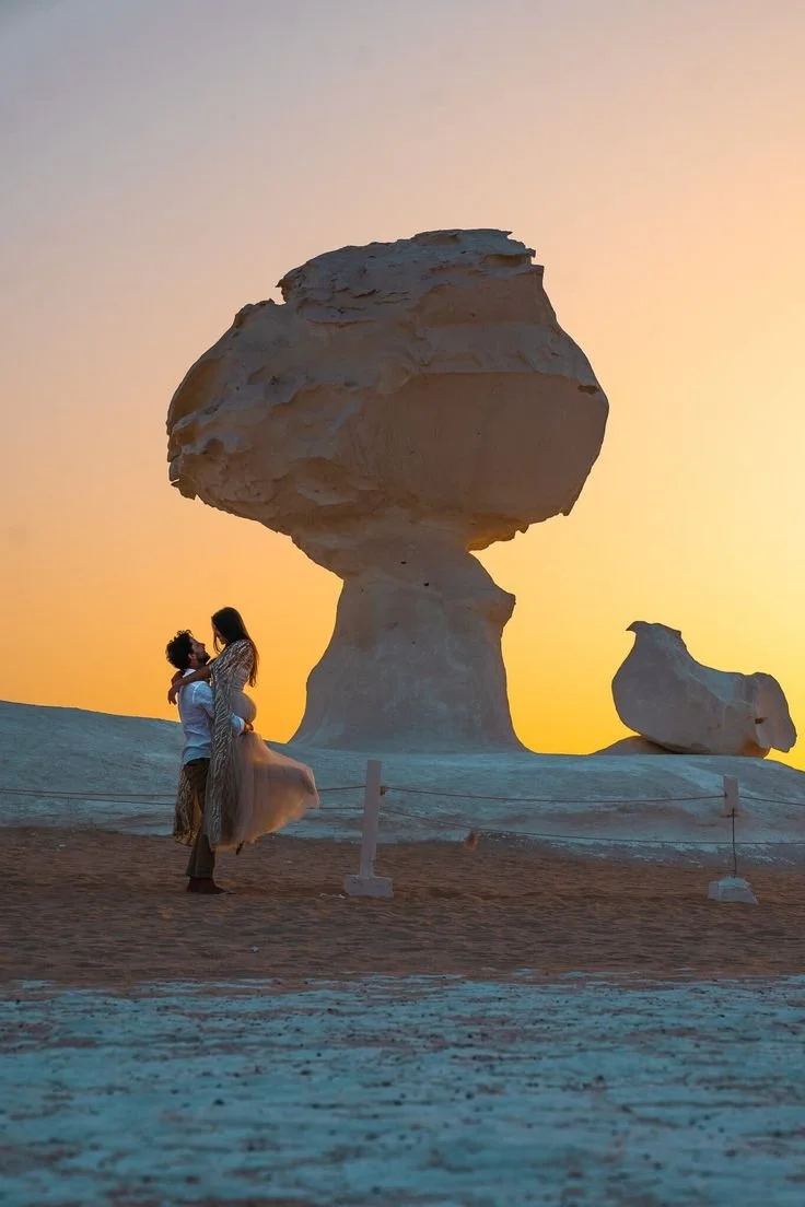 A couple embraces in a desert landscape at sunset with large, unusual rock formations in the background.