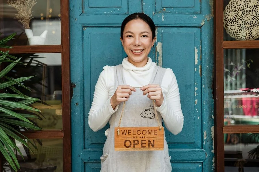 photo of woman standing in front of blue door smiling and holding an open sign