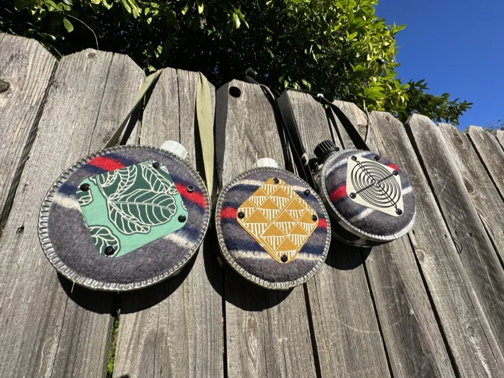 Three decorated metal canteens hanging on a wooden fence; each covered with patterned fabric and fitted with a strap.