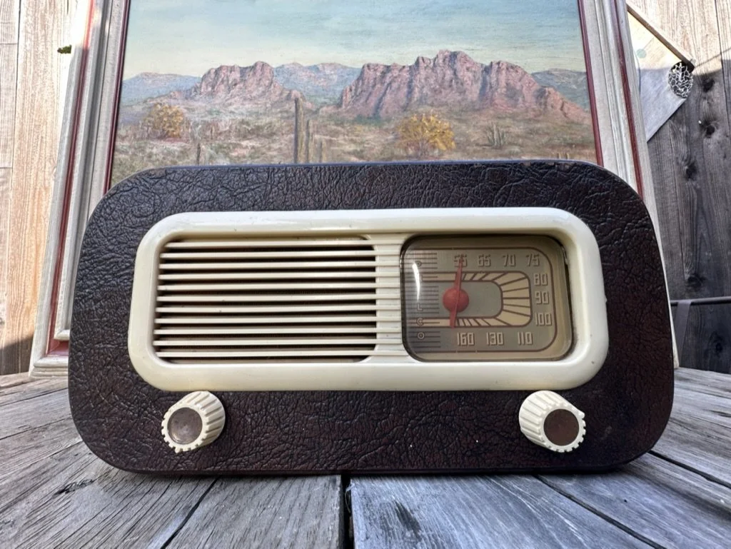 Vintage radio with brown casing and two dials, placed on wooden surface with a landscape painting in the background.