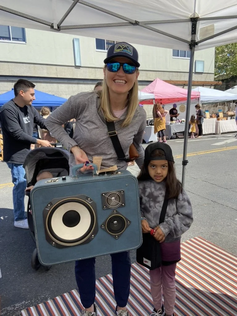 A woman and a child standing under a market tent, the woman is holding a vintage suitcase with speakers attached, resembling a portable sound system. They are both smiling and wearing casual outdoor clothing at a street market.