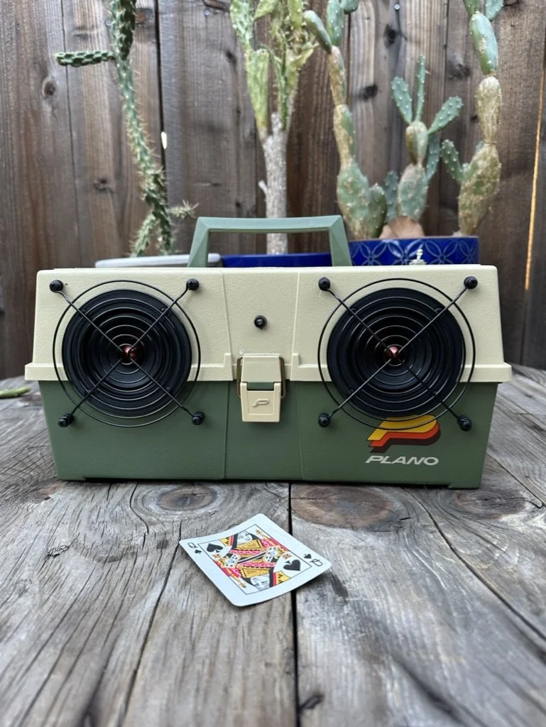 A vintage boombox resembling a green and beige fishing tackle box with the logo "Plano." It has large black speaker grills with a handle on top, set on a wooden surface beside a queen of spades playing card. Cacti are in the background against a wood