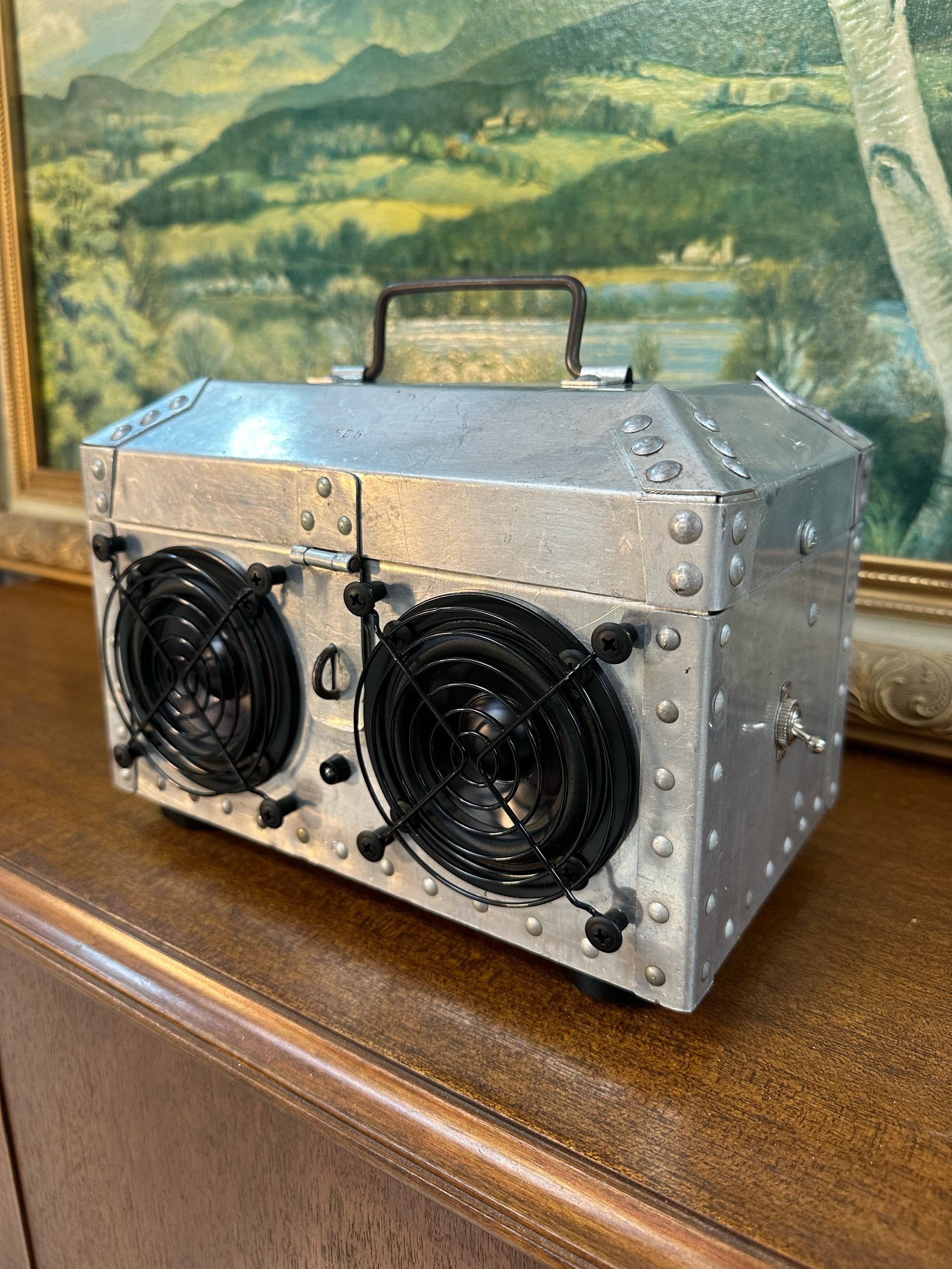 Metallic box with two cooling fans and a top handle, placed on a wooden surface. A landscape painting is in the background.